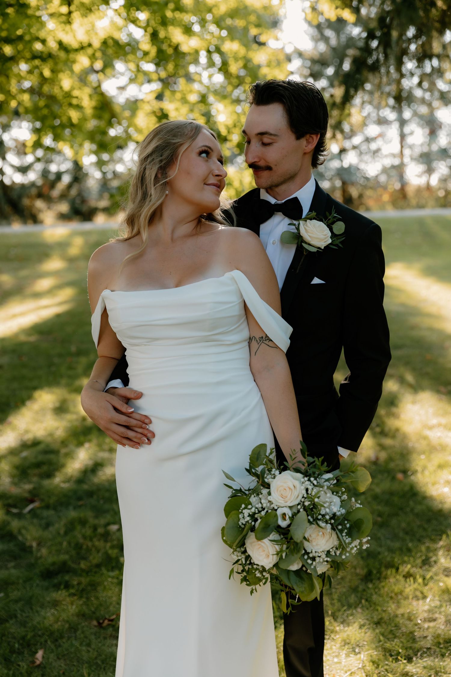 Bride in off-shoulder white gown and groom in black tuxedo sharing romantic glance at outdoor wedding.