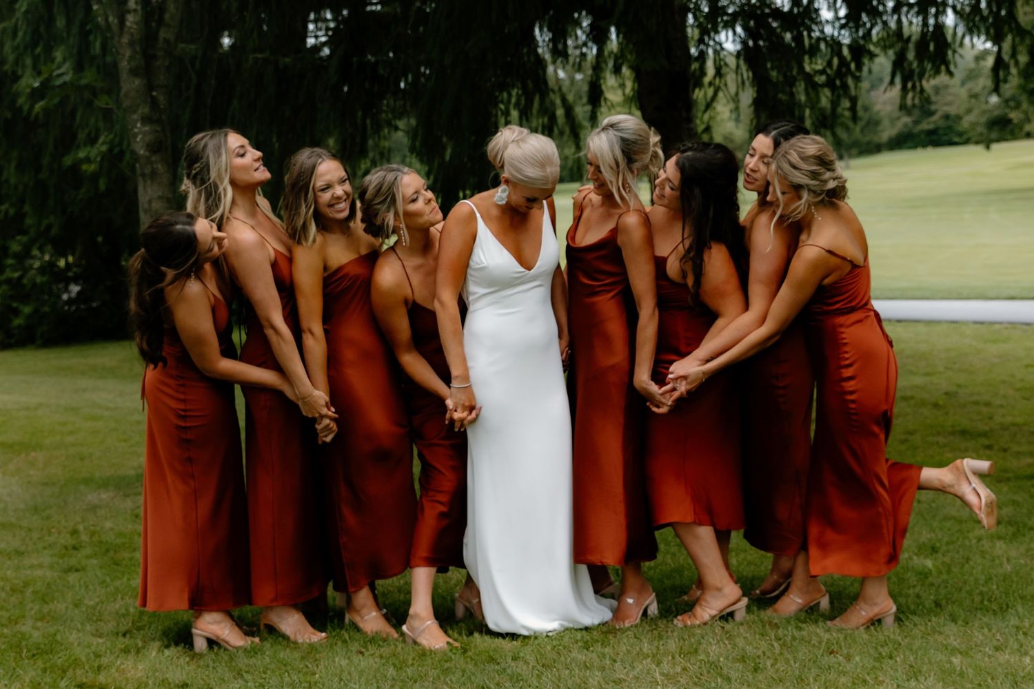 Bridesmaids in rust satin dresses surrounding bride in white gown holding hands on green lawn.
