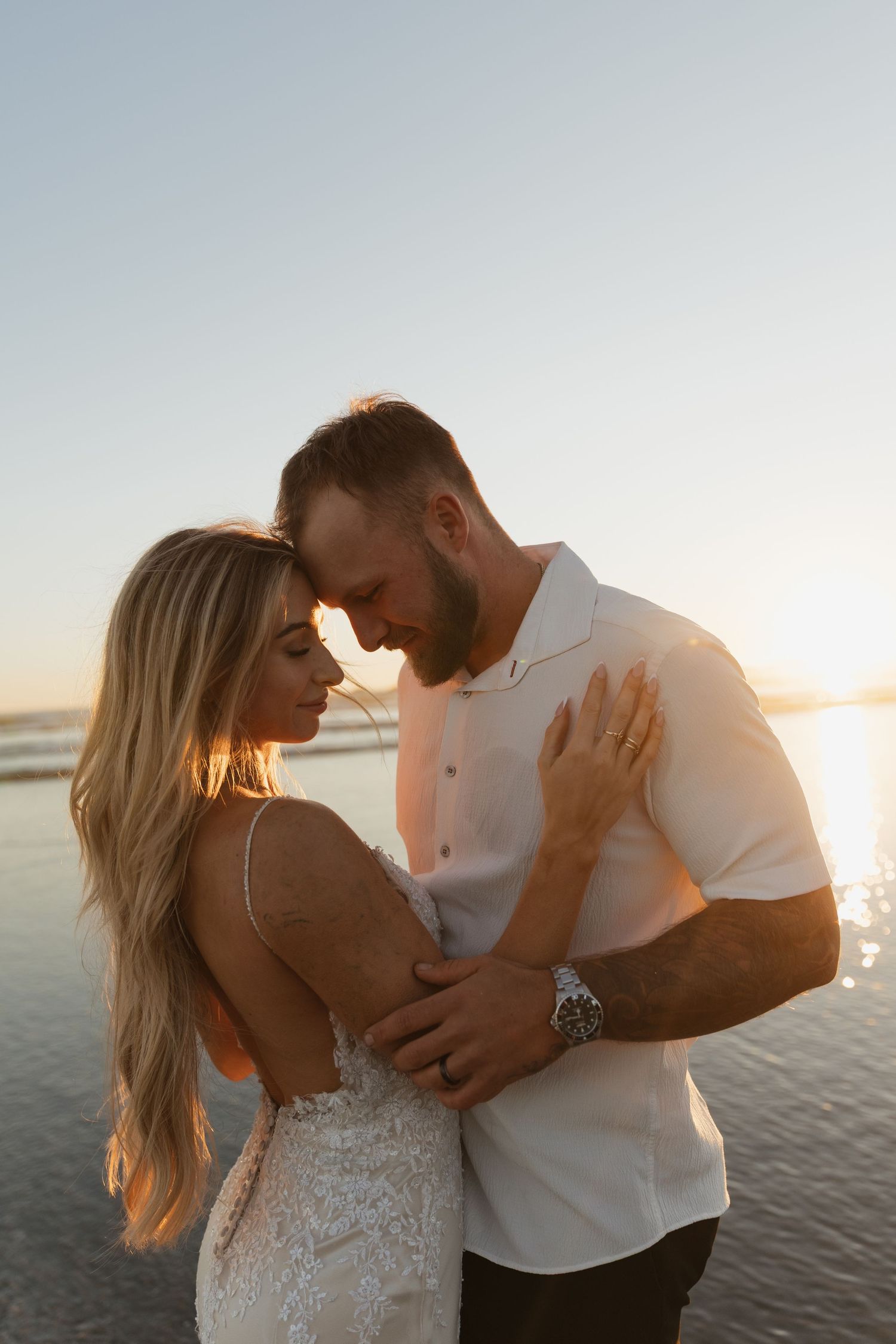 Couple embraces at golden hour on a beach, glowing in warm sunset light near the ocean.
