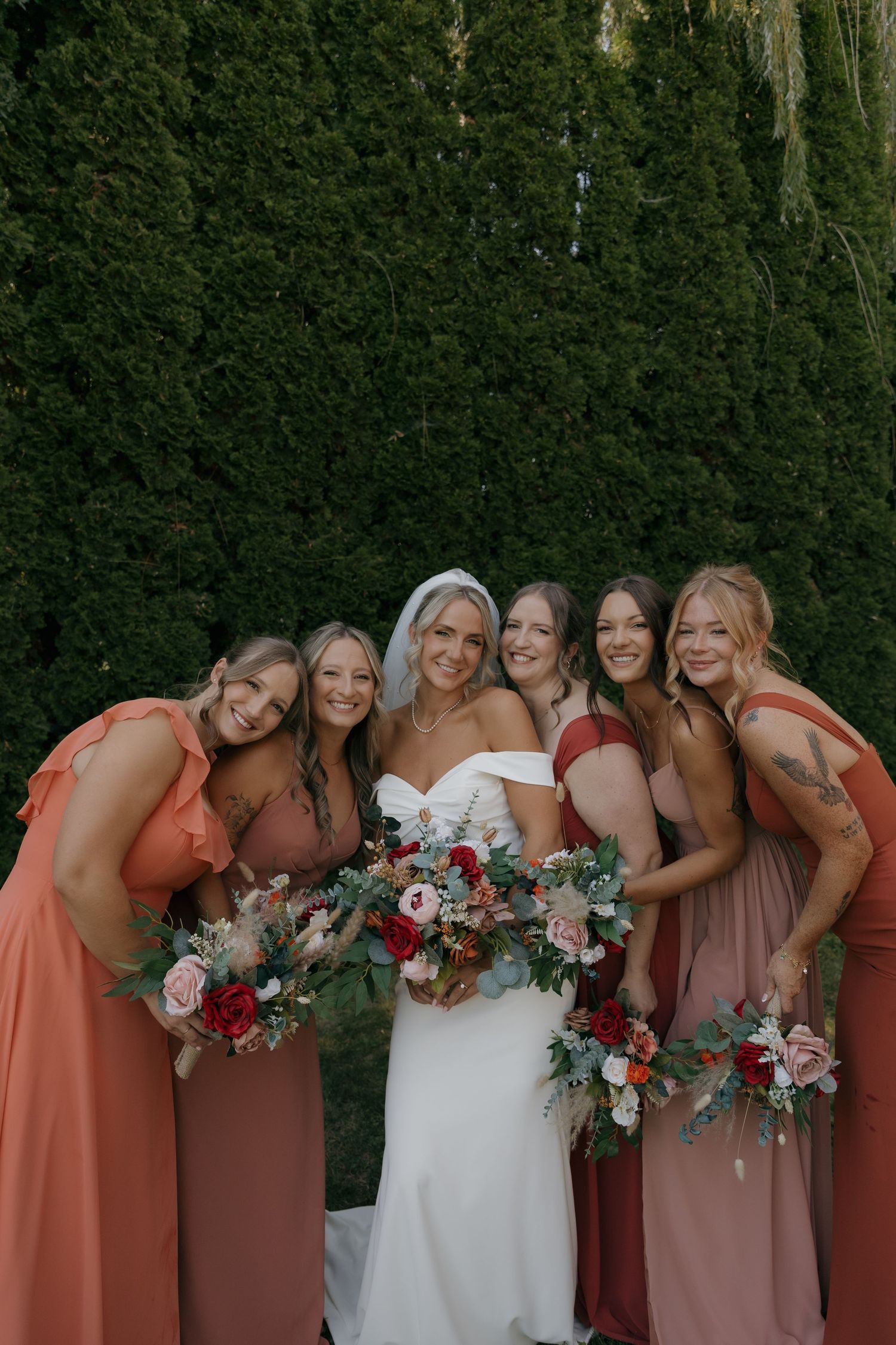 Smiling bride and five bridesmaids in rust and mauve dresses pose with lush floral bouquets.
