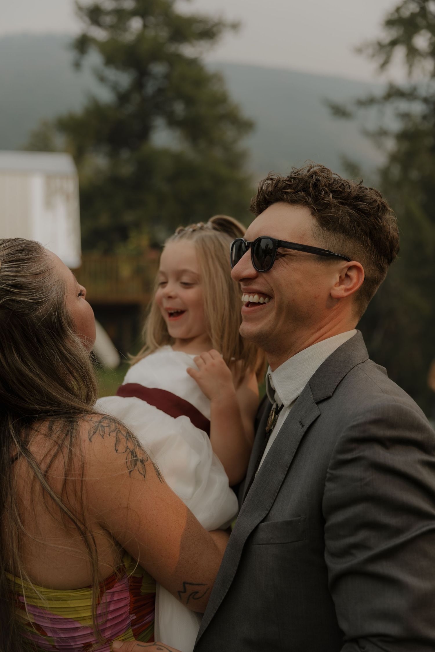 Man in gray suit laughing joyfully while holding a young girl at outdoor wedding.