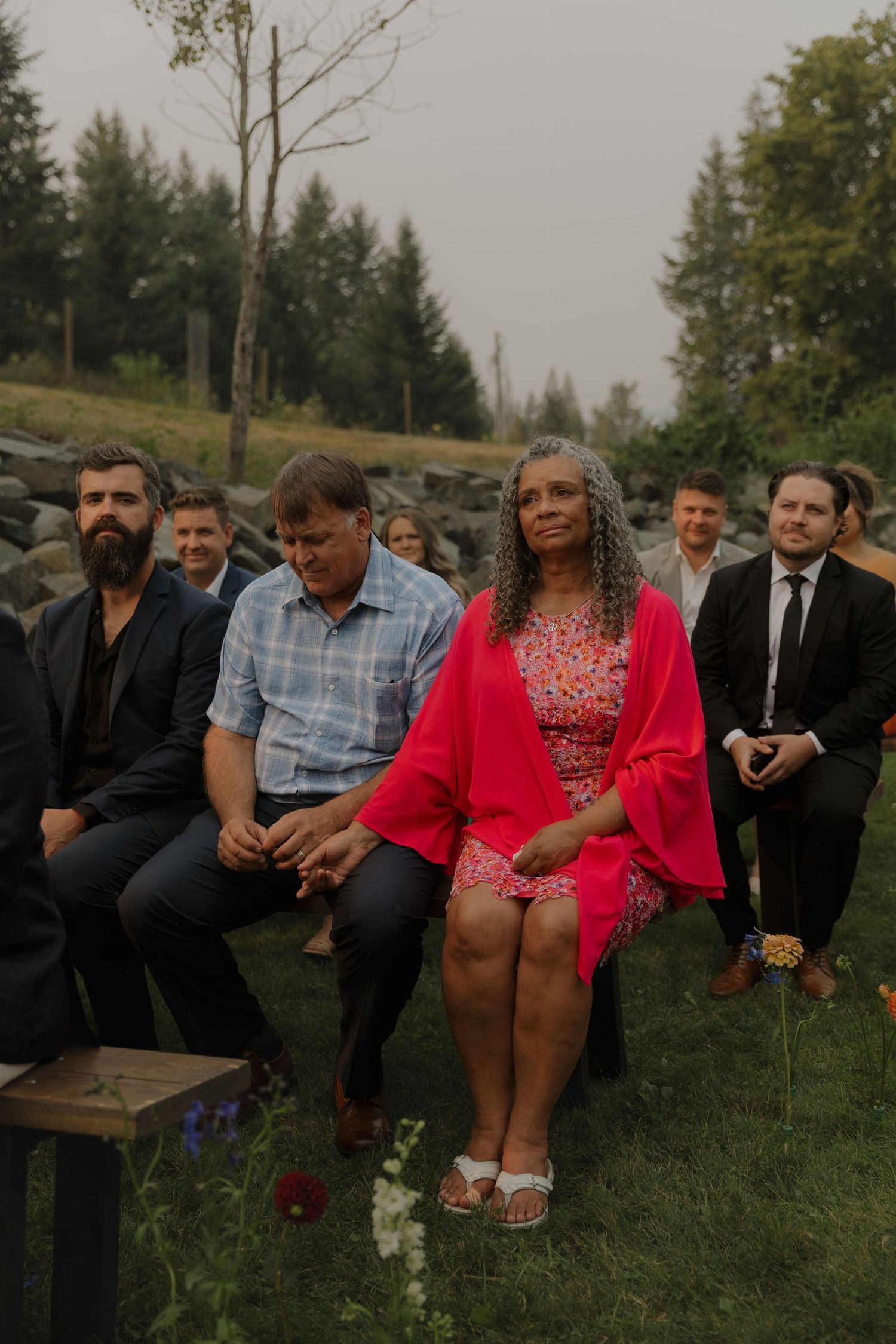 Wedding guests seated outdoors watching ceremony, woman in red dress standing prominently.