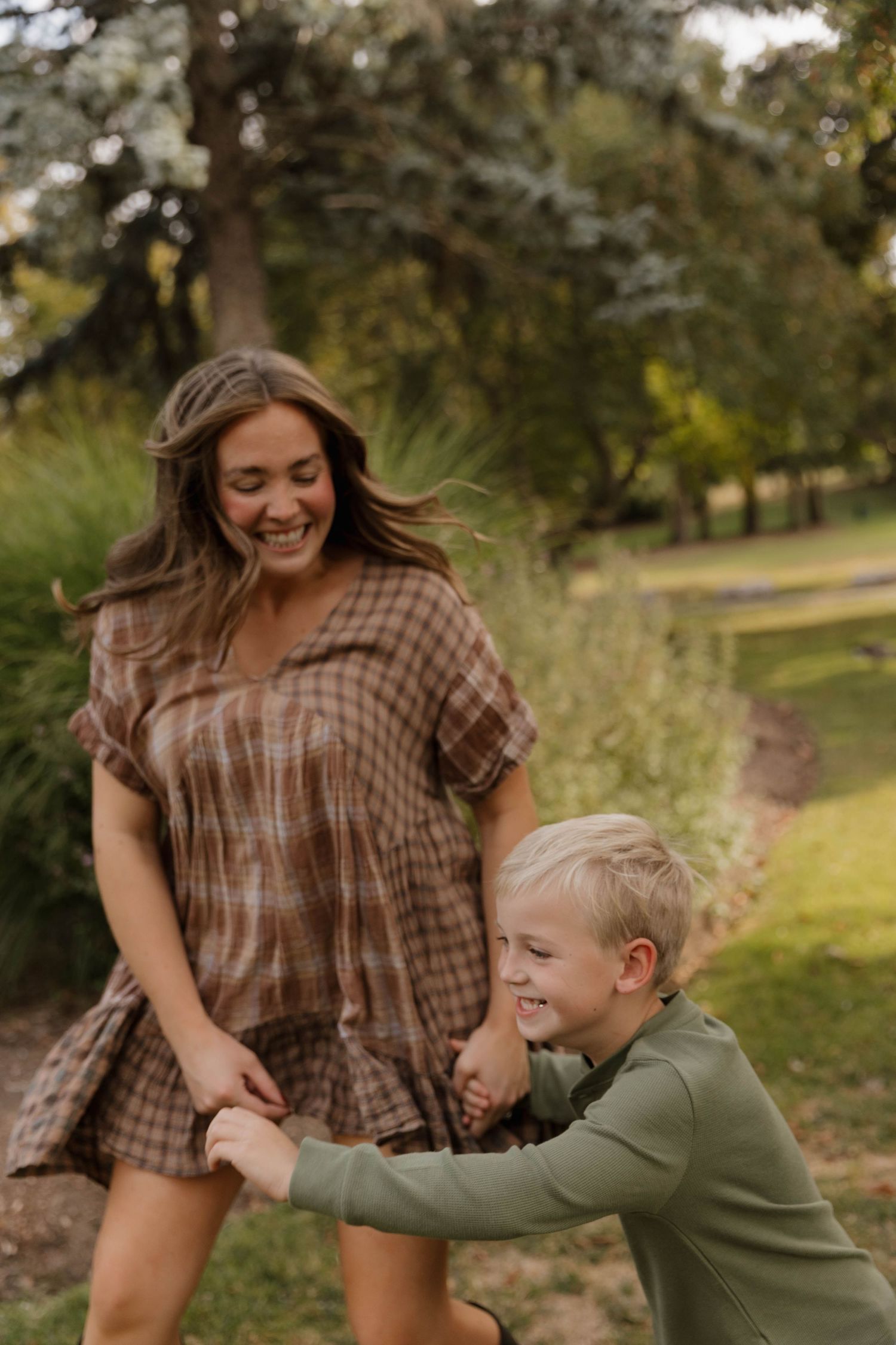 Joyful mother in plaid dress spinning with young blonde son in green park.