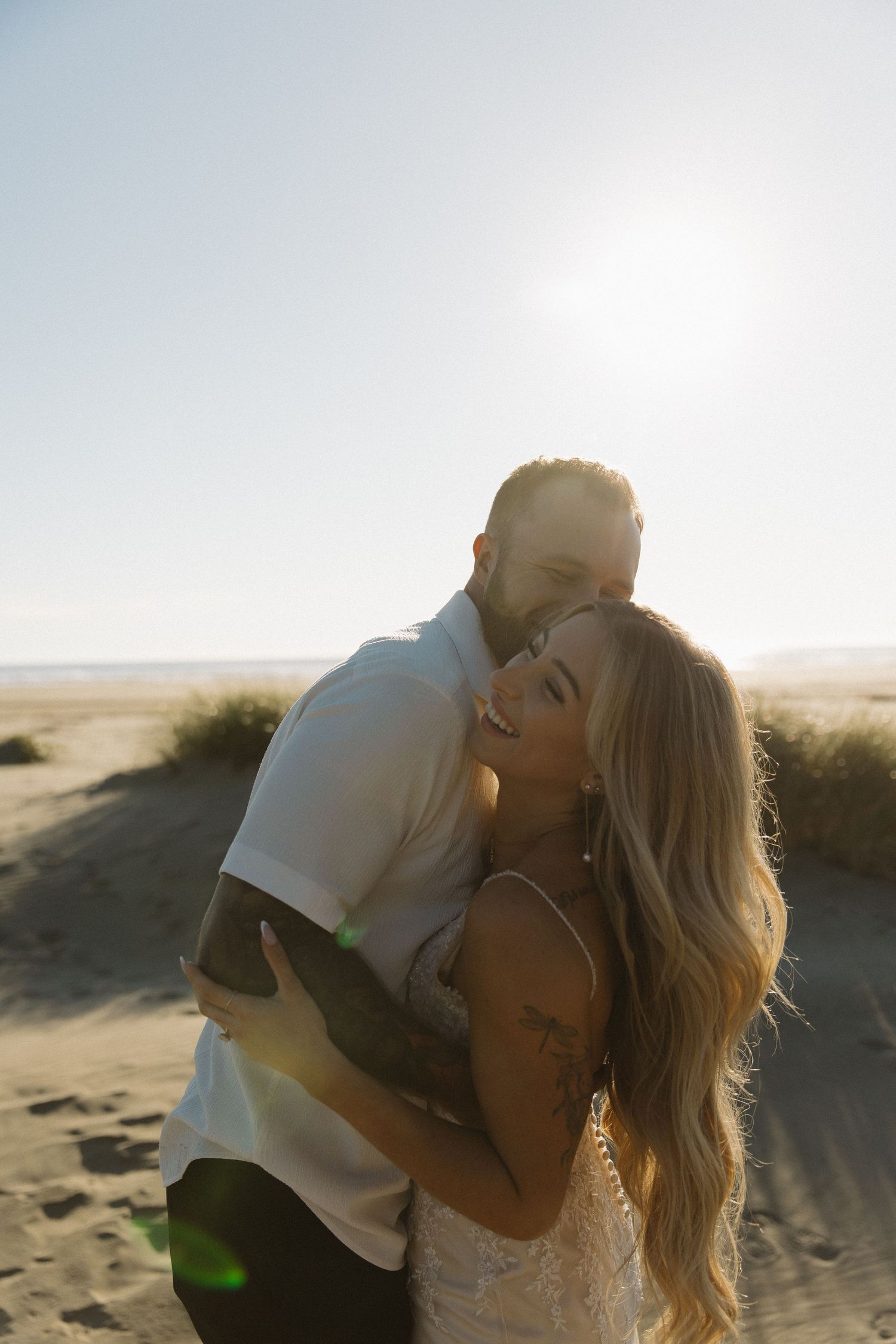 Newlyweds embracing on sandy beach with bright sun flare in clear sky.