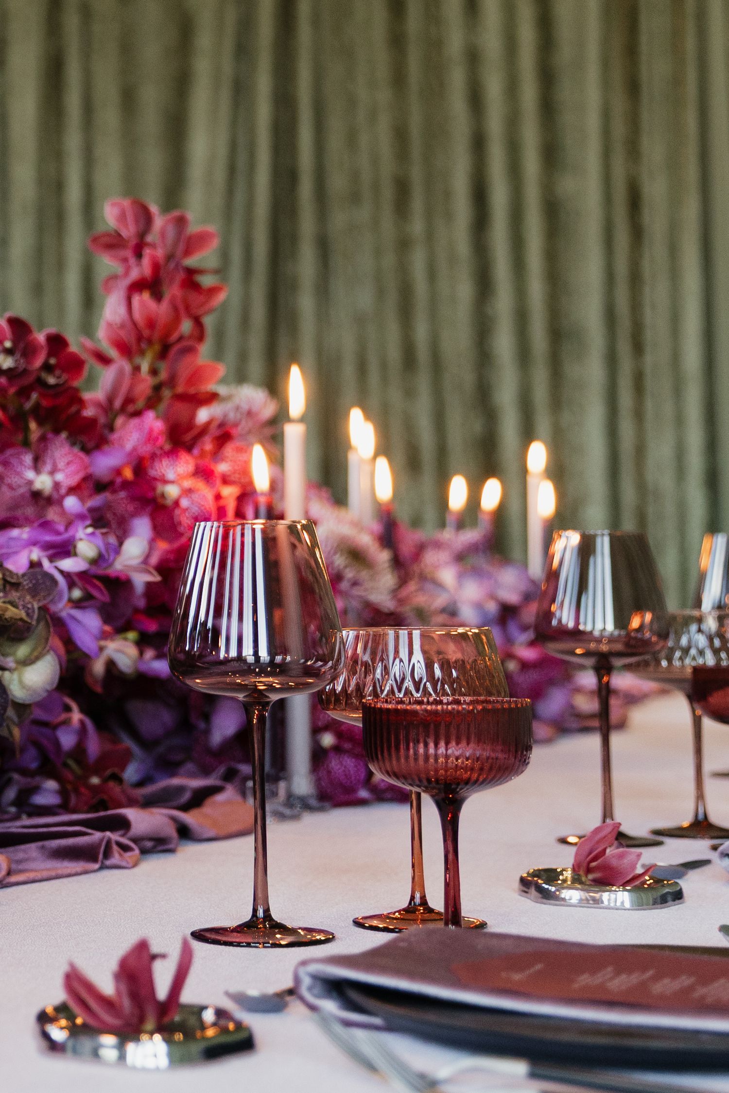 Wedding reception table detail with mauve tinted glassware, lit candles, and purple floral arrangements.