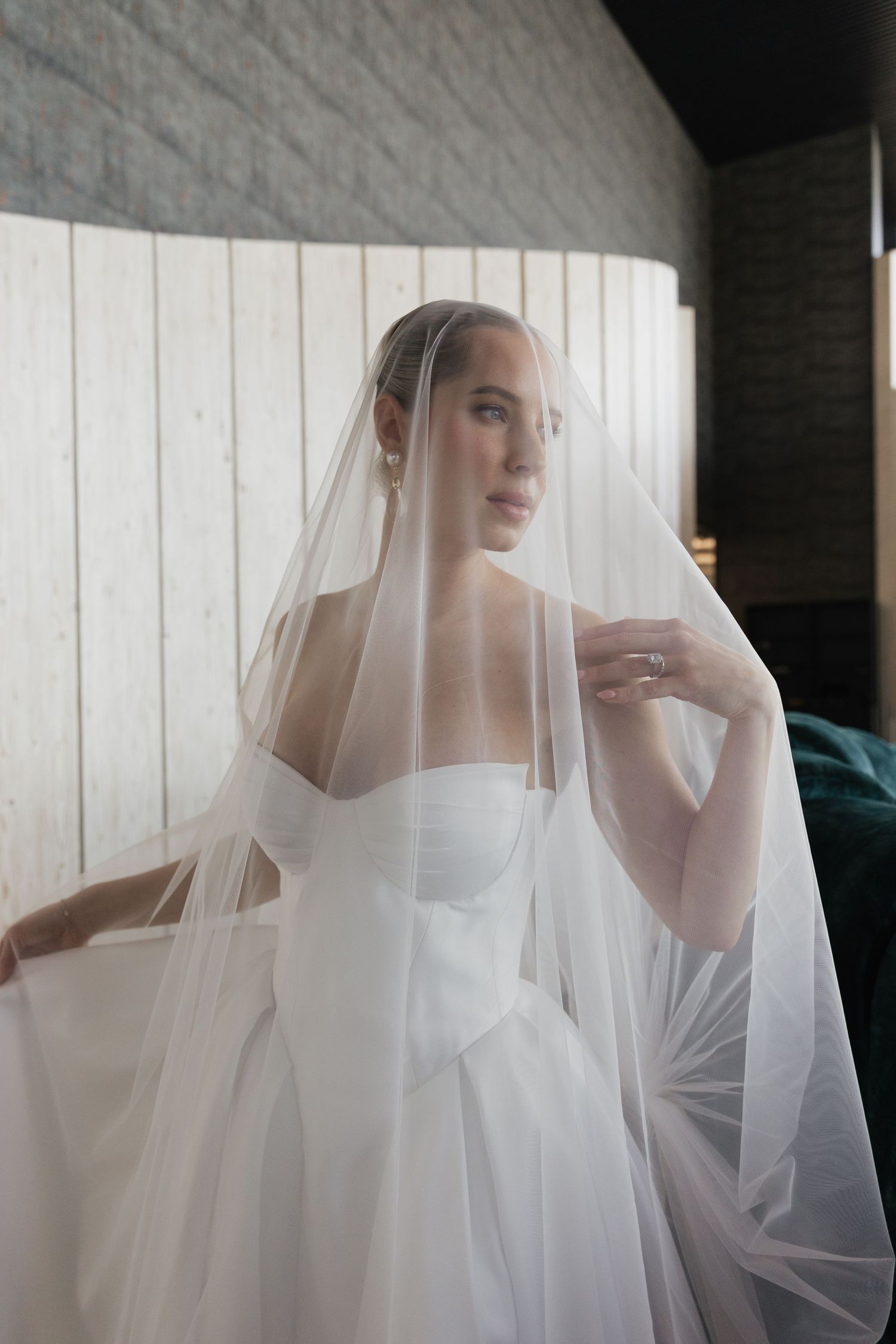 Beautiful bride in a white strapless gown lifts her flowing veil in a softly lit indoor setting.