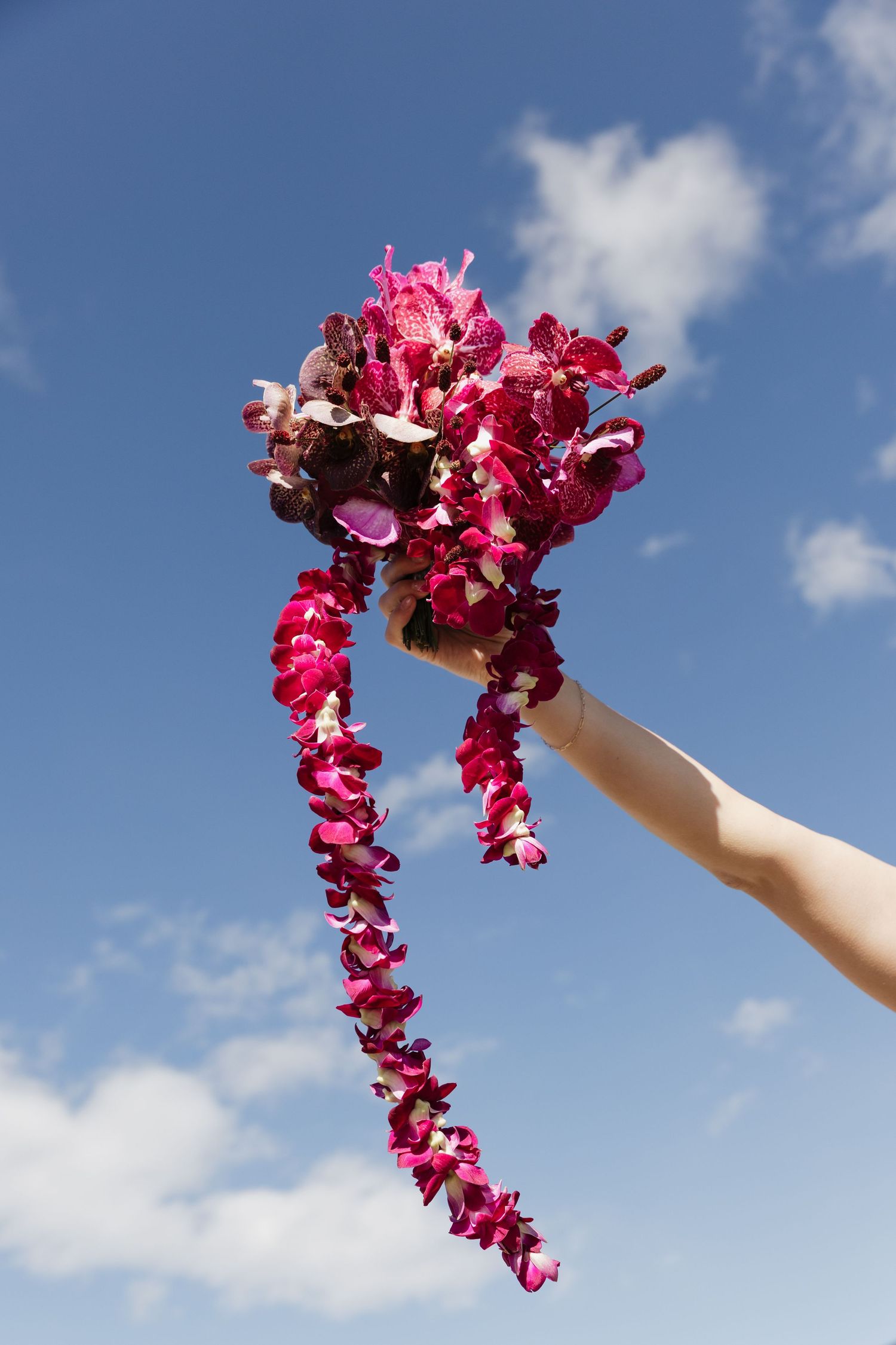 Hand holds a cascading bridal bouquet of vibrant pink and burgundy orchids against a blue sky.