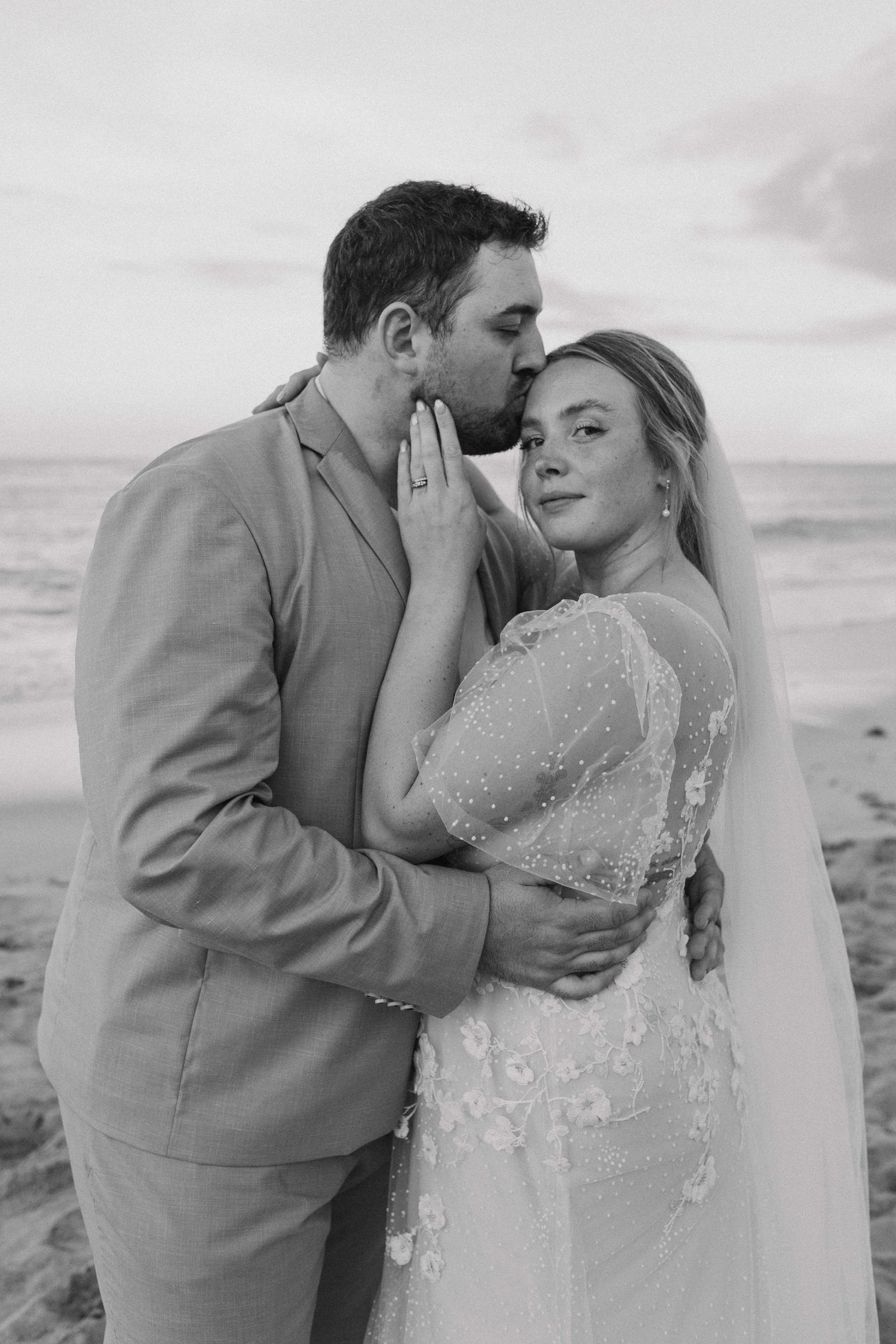 Black and white photo of groom kissing bride on forehead at beach wedding.