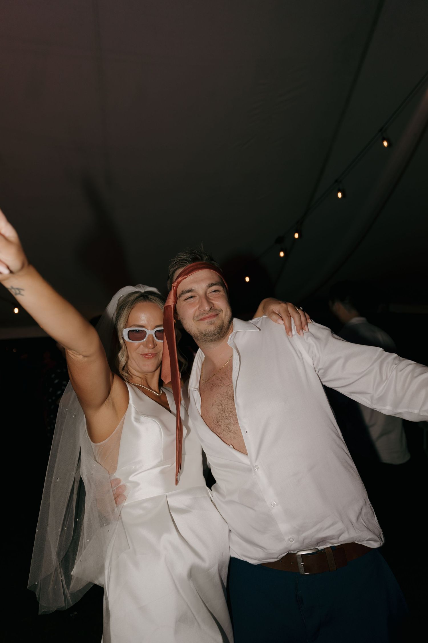 Bride in white gown and sunglasses posing with guest at lively wedding reception.