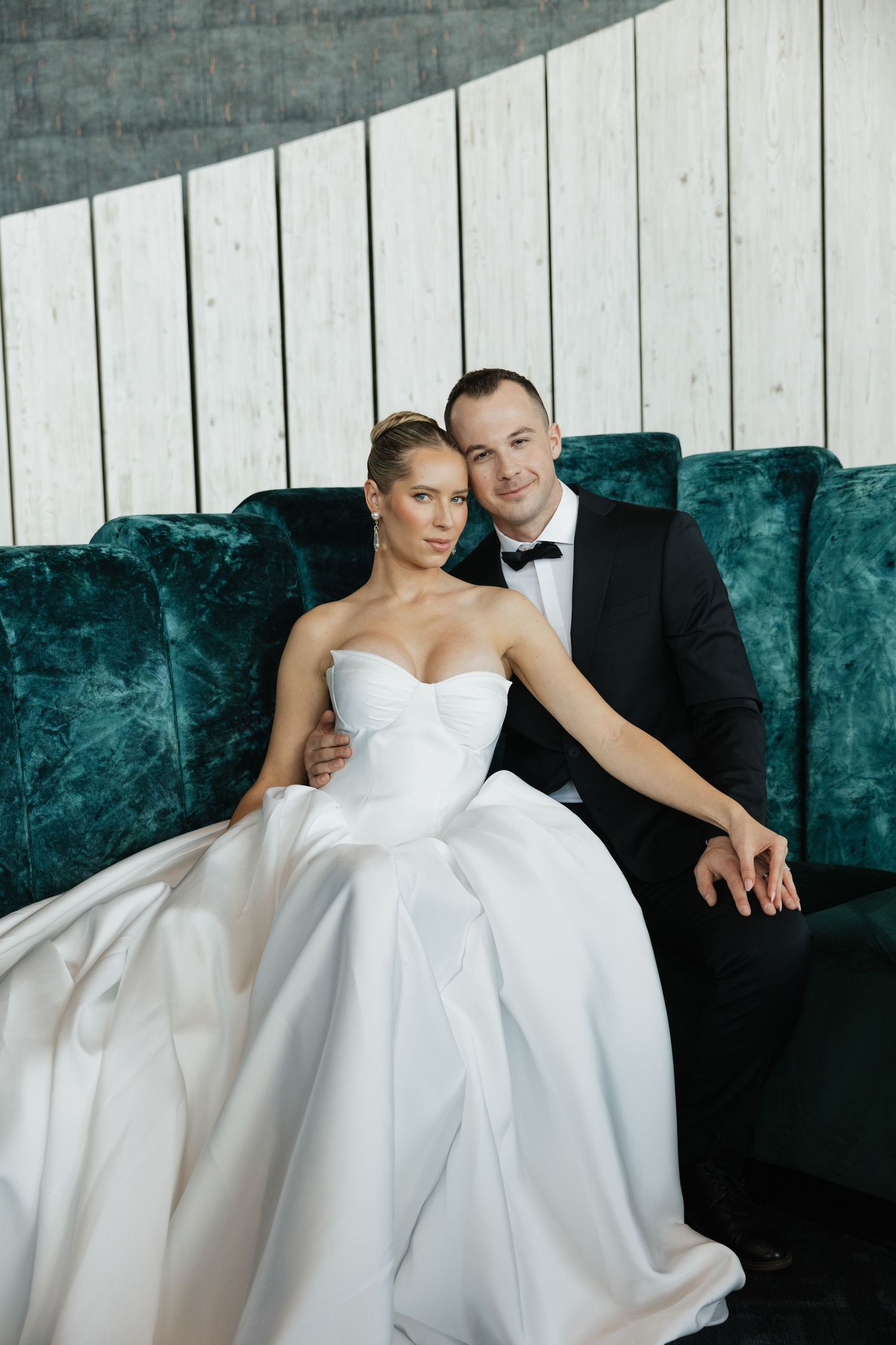Bride and groom pose together on teal velvet sofa, she in white ball gown, he in classic black tuxedo.