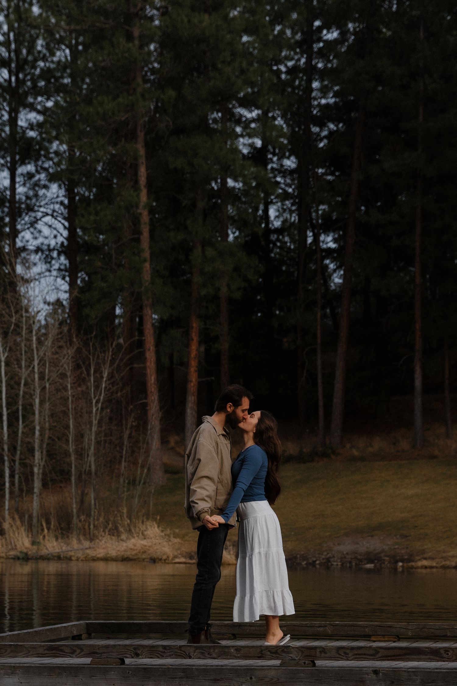 Couple sharing a kiss on a lakeside dock with tall pine trees in the background at dusk.