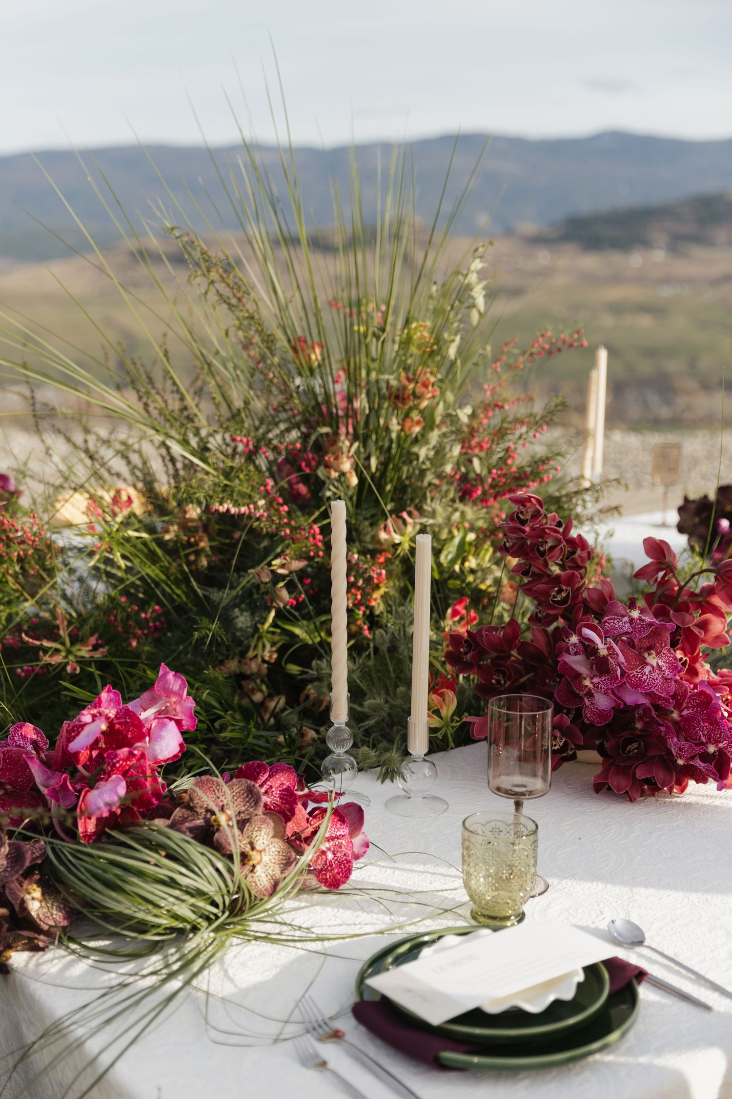 Luxurious outdoor wedding tablescape with vibrant pink and burgundy orchids, tall candles, and mountain views.