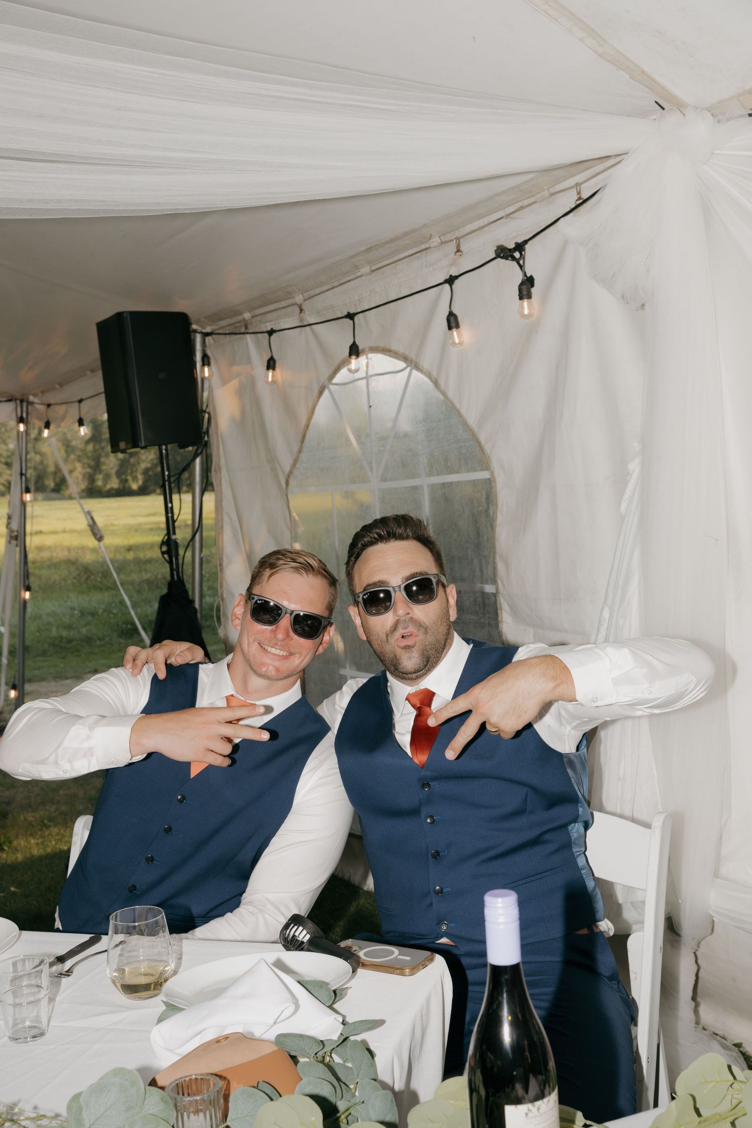 Two groomsmen in navy vests and sunglasses posing playfully at wedding reception table.