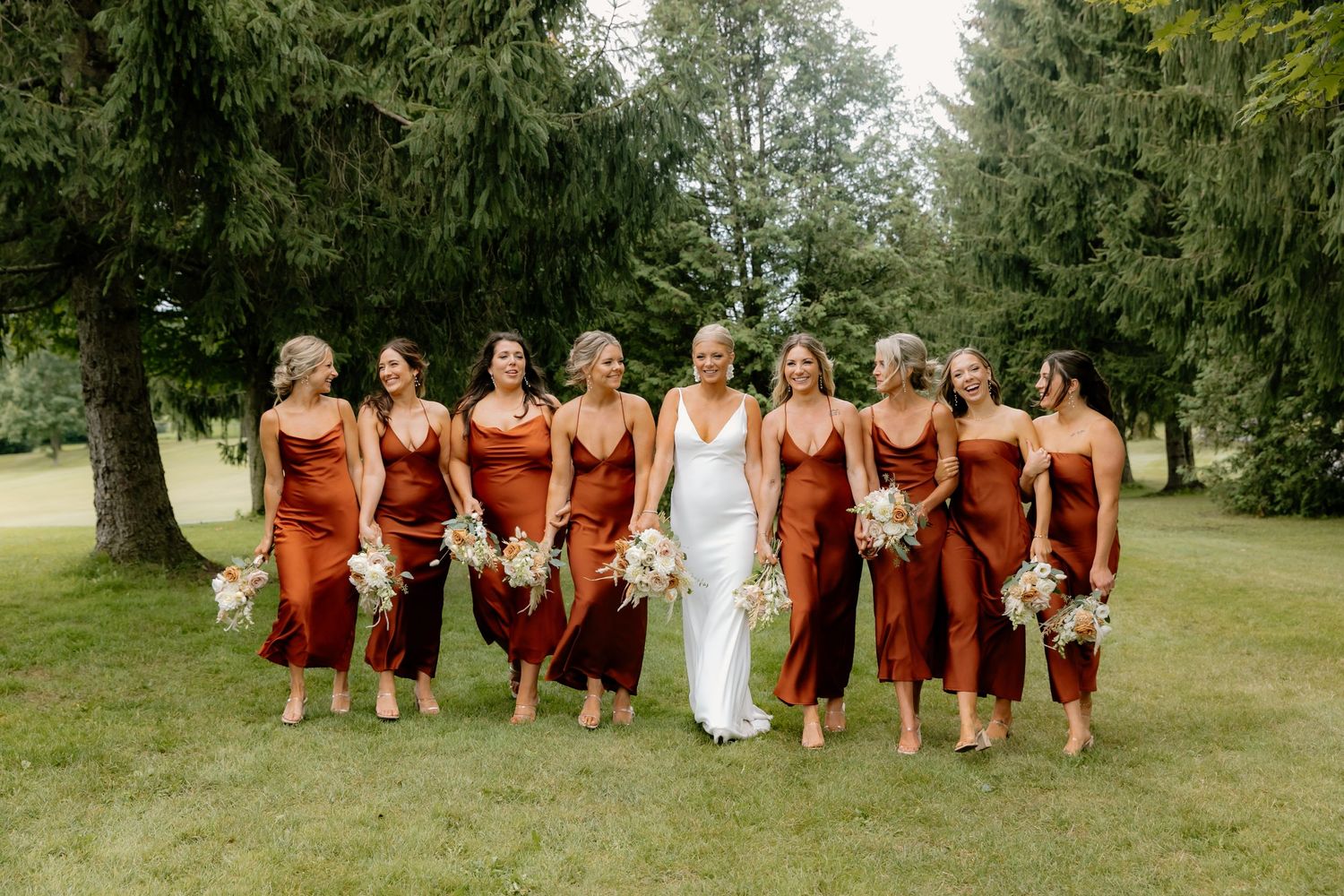 Bride in white gown walks with bridesmaids in rust-colored satin dresses holding bouquets on green lawn.