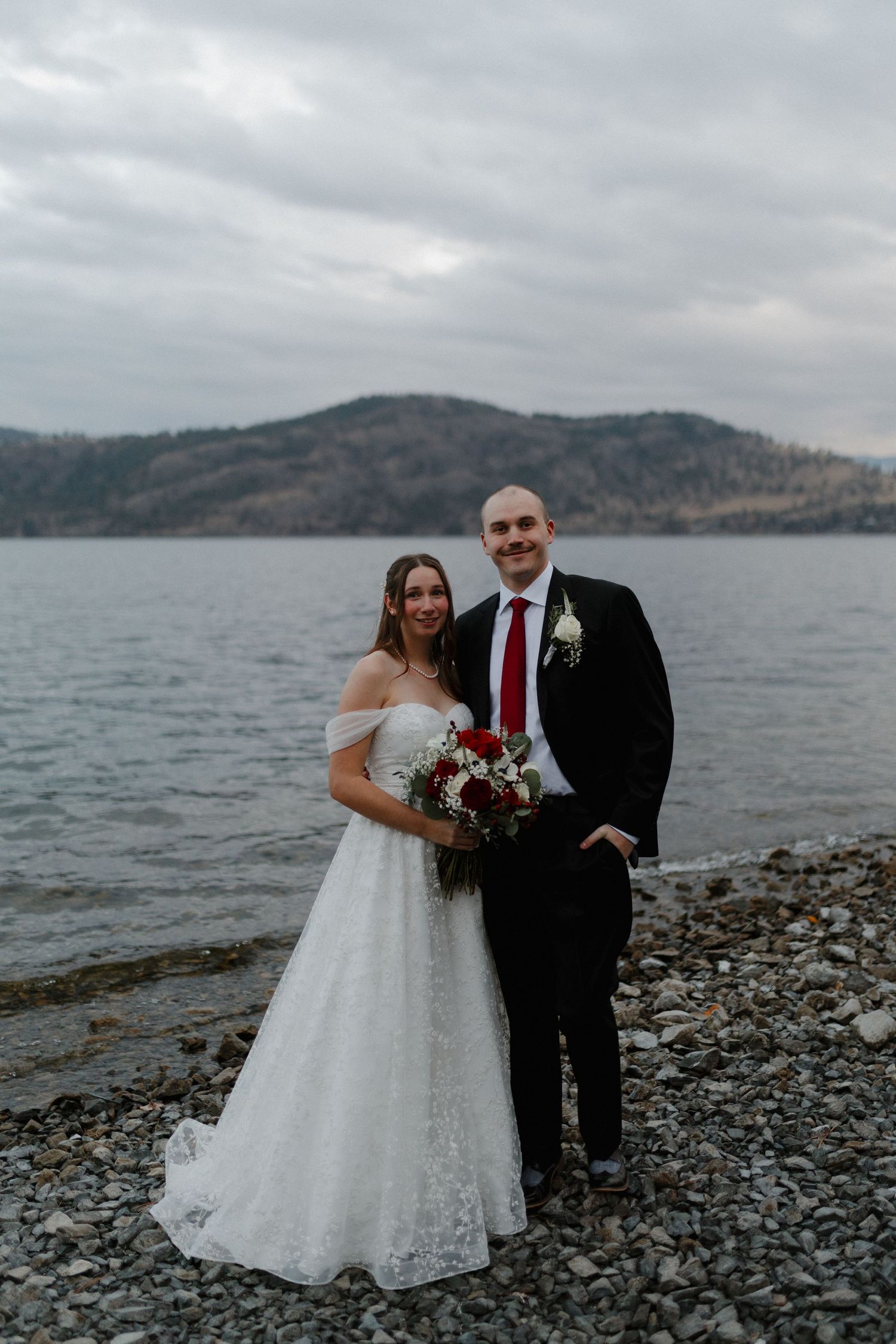 Bride and groom posing by rocky lakeside with mountain backdrop on cloudy day.