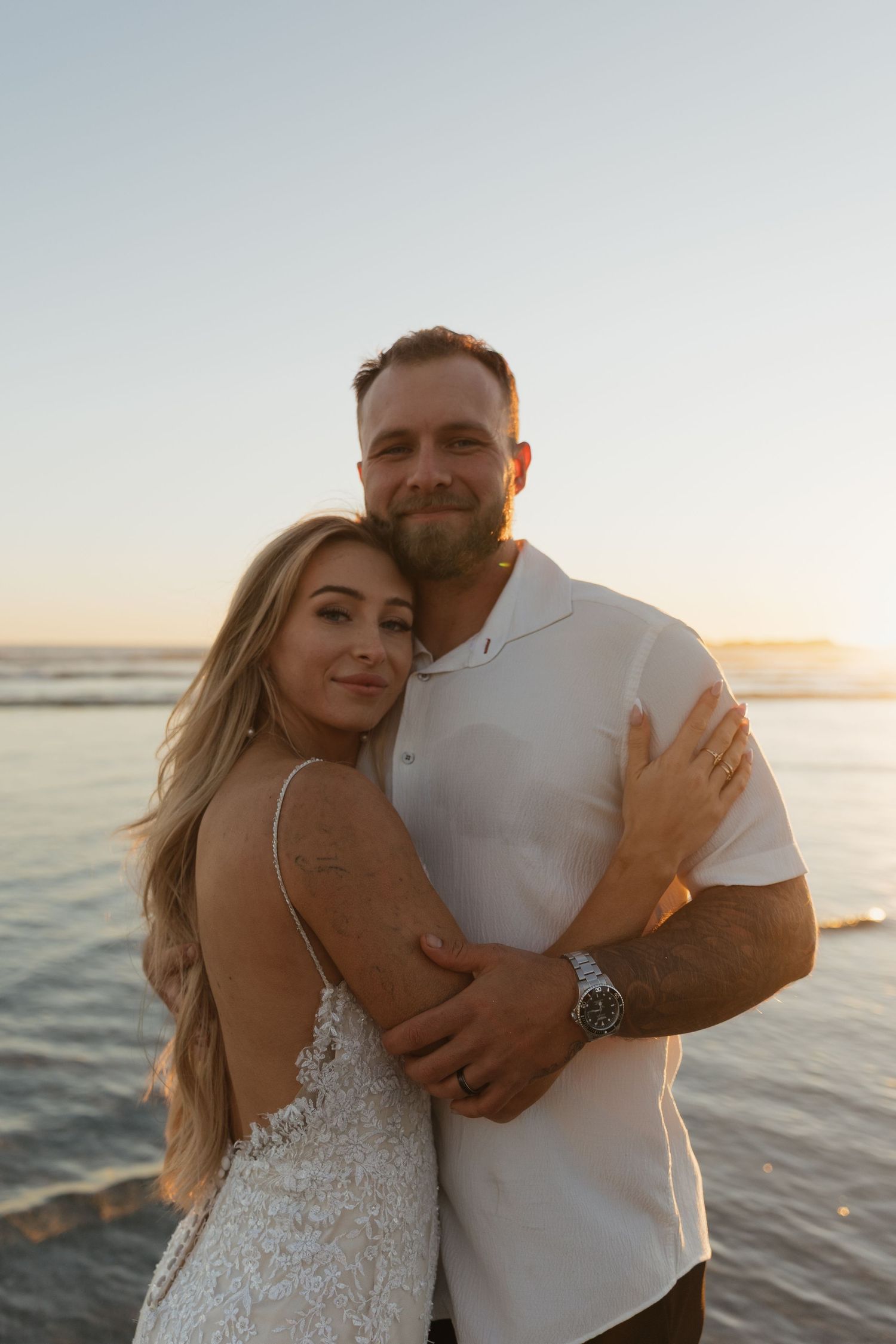 Smiling couple embraces at sunset by the ocean, bride in lace gown and groom in white shirt.