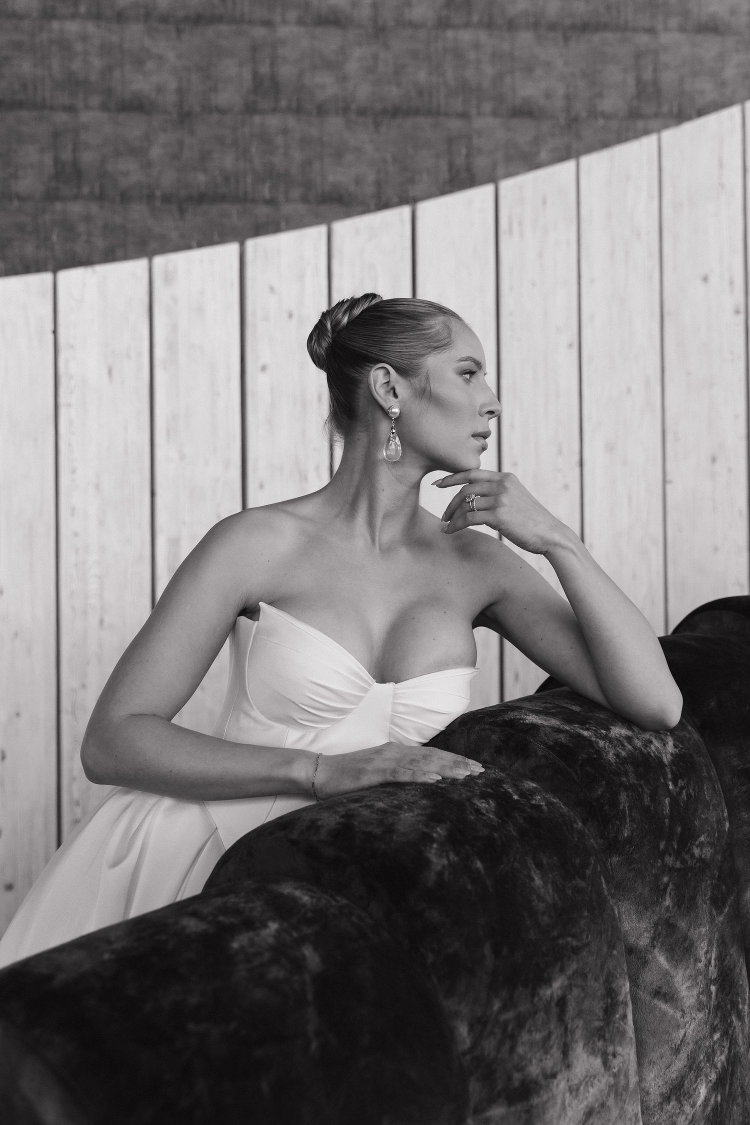 Black and white portrait of bride in strapless wedding dress leaning on velvet sofa with wooden wall backdrop.