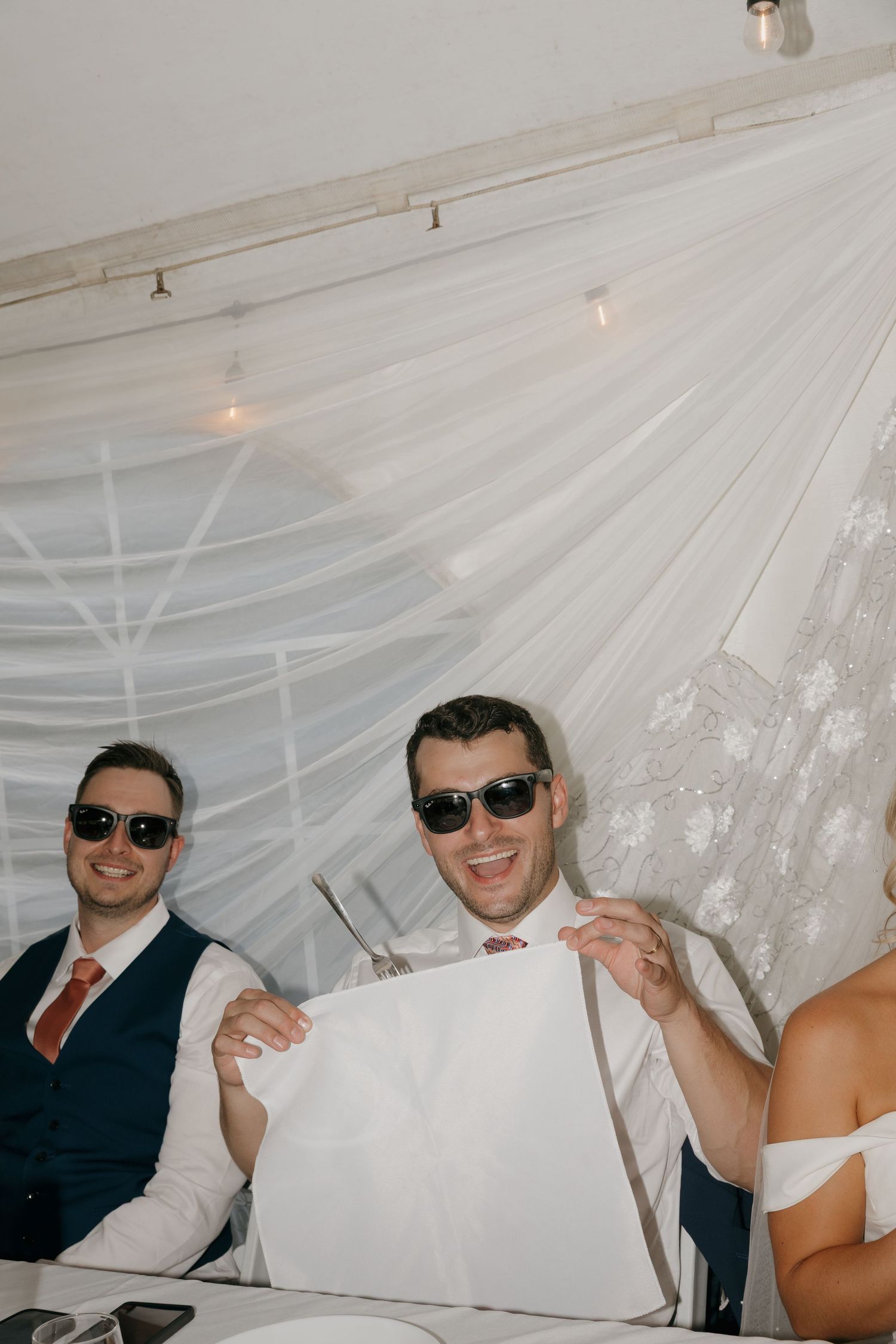Two groomsmen in navy vests wearing sunglasses holding white gift bag at wedding reception.