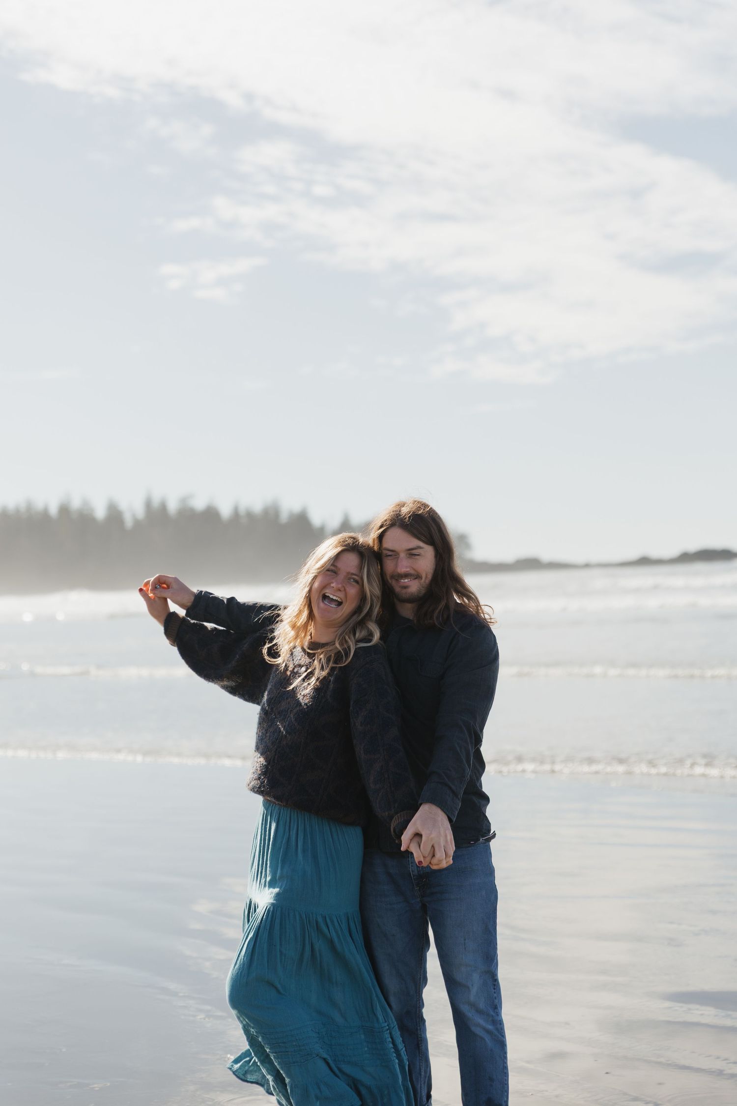 A happy couple dancing and laughing together on a scenic beach with ocean waves behind them.