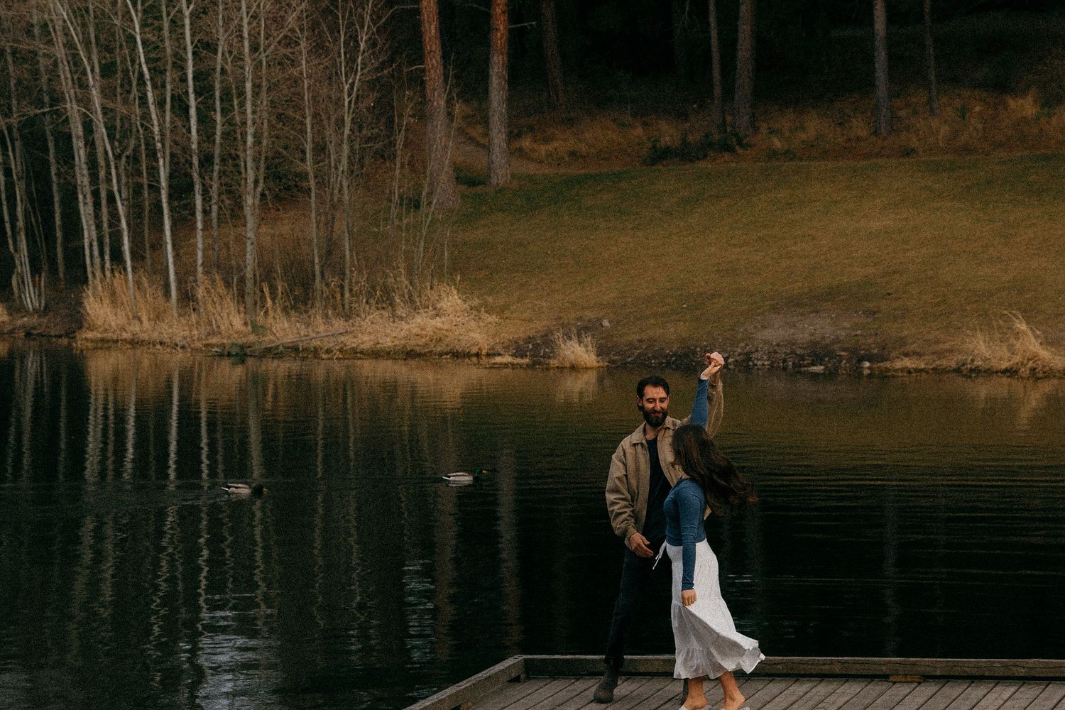 Couple dancing on a wooden dock by a calm reflective lake surrounded by autumn trees.