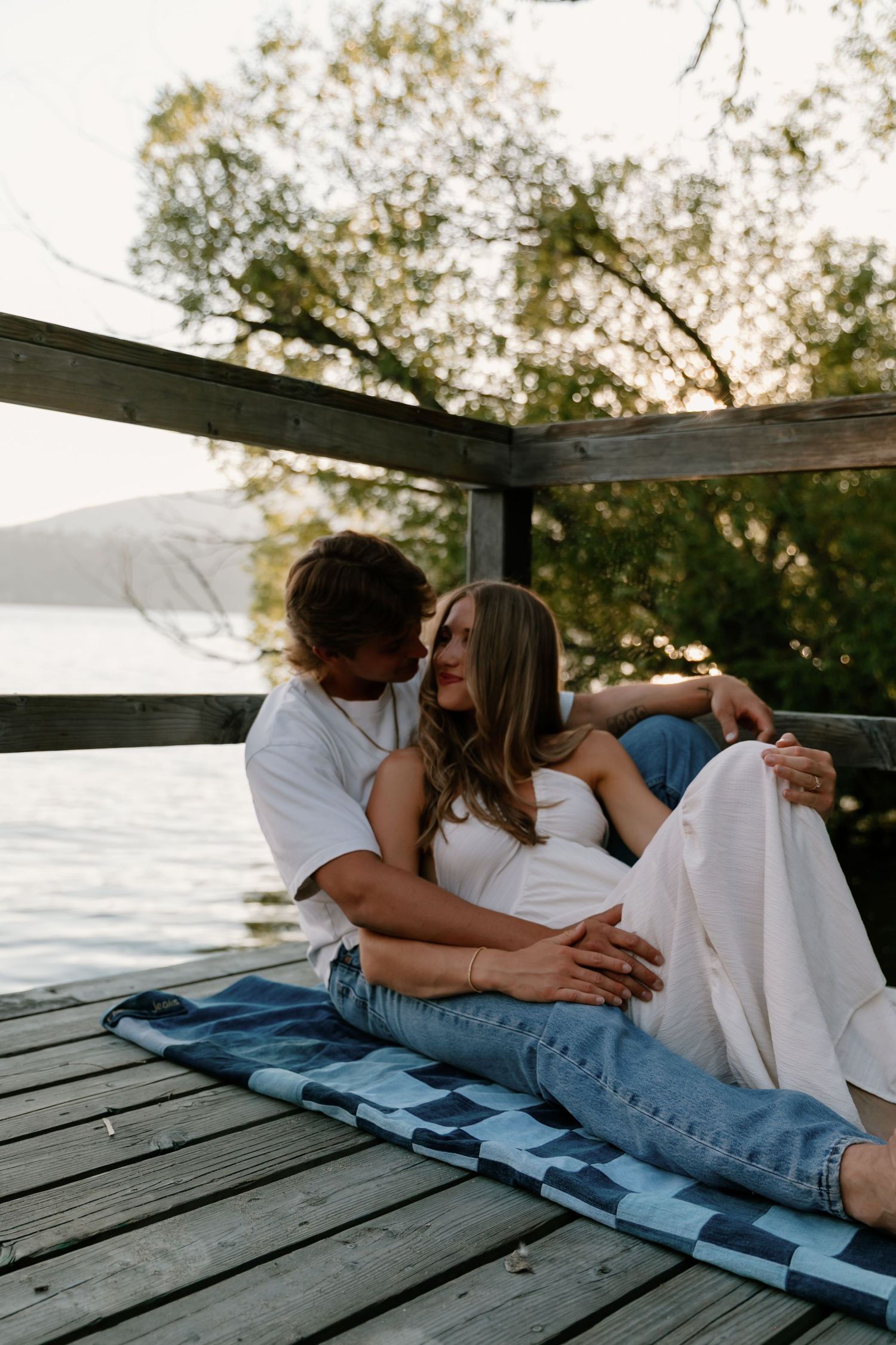 Couple cuddling on a wooden lakeside dock at sunset, woman resting against man with lake views behind.