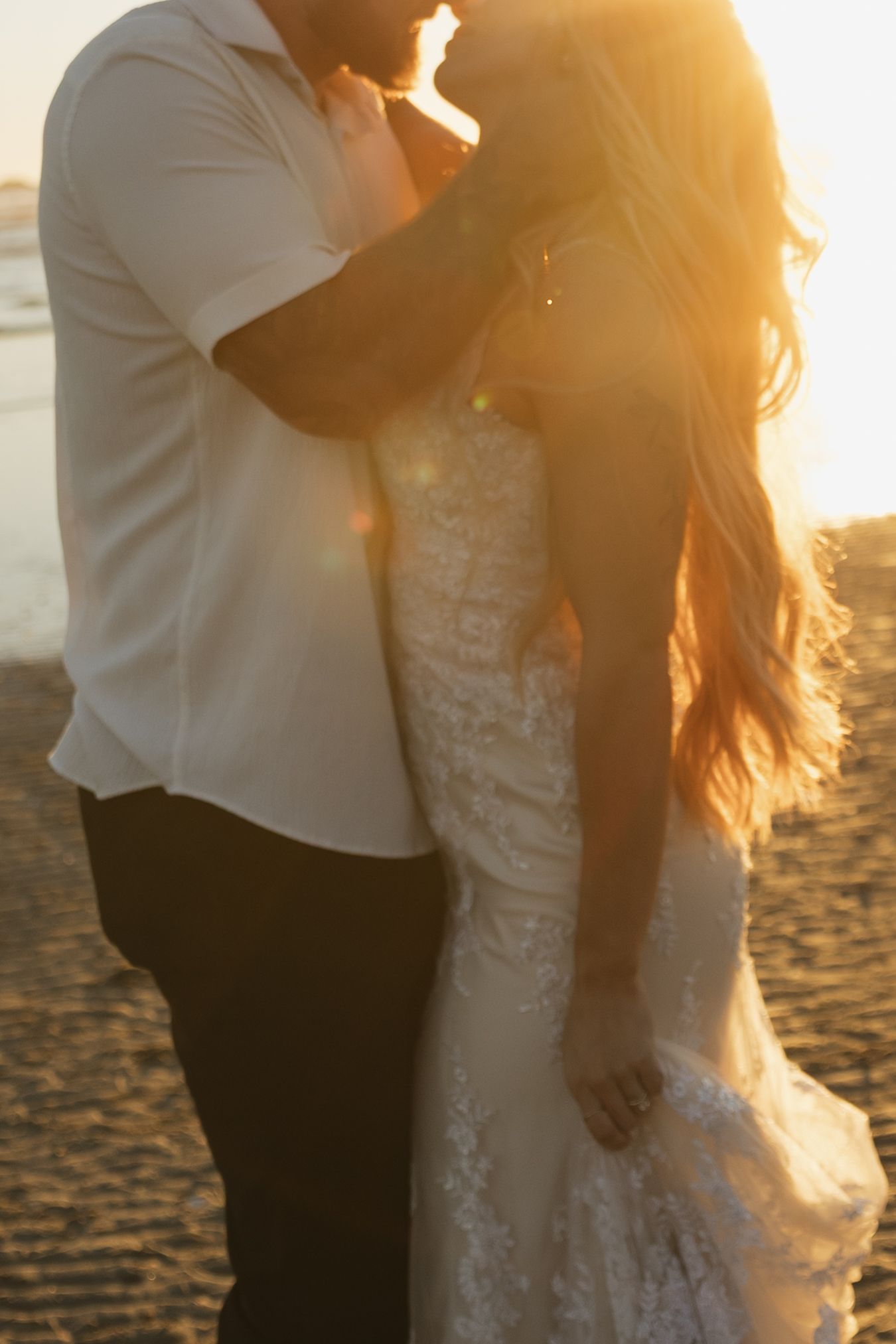 Couple embraces in a golden sunset glow on the beach, bride in intricate lace wedding gown.