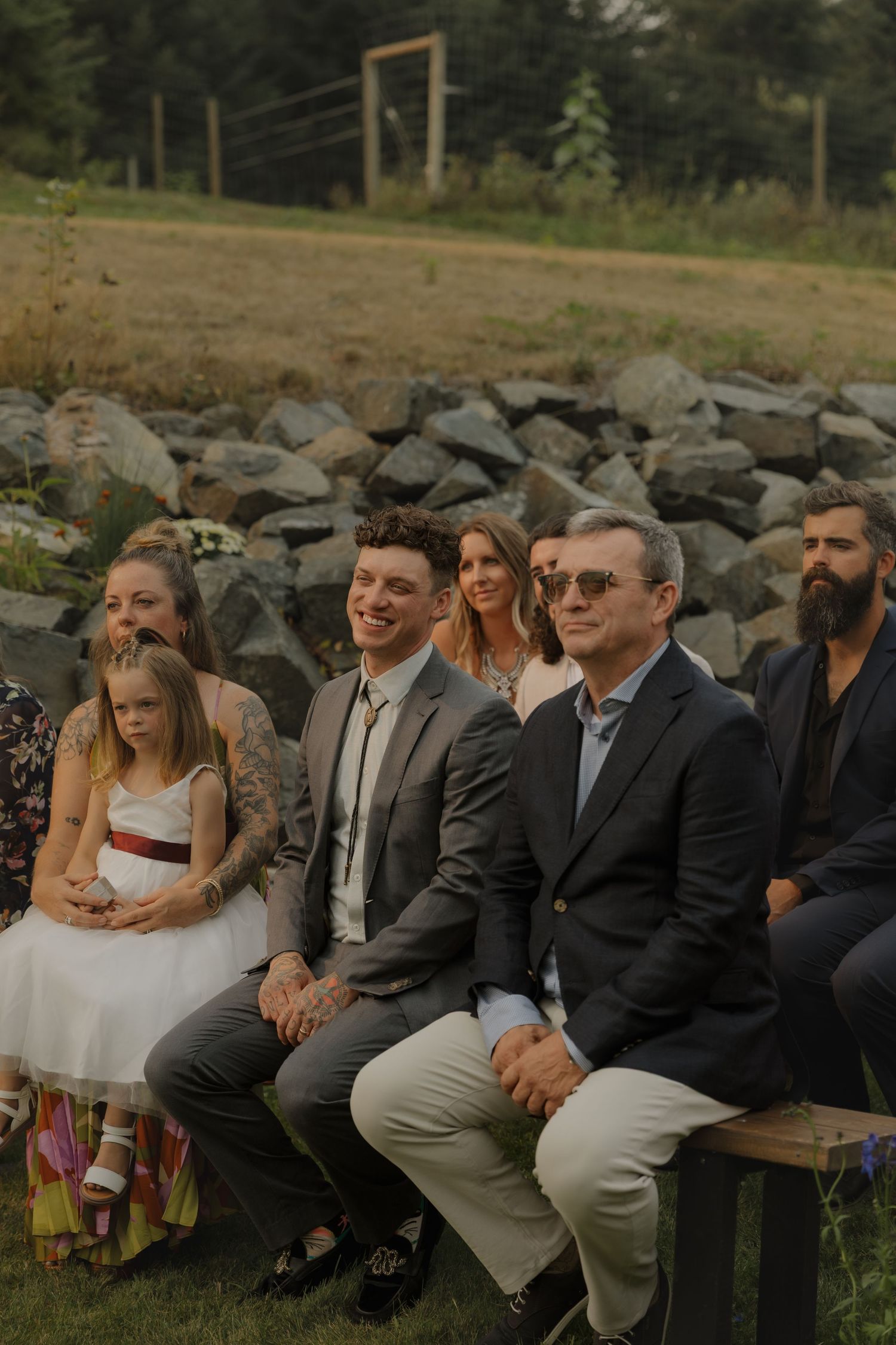 Wedding guests seated outdoors smiling during ceremony, rocky landscape in background.