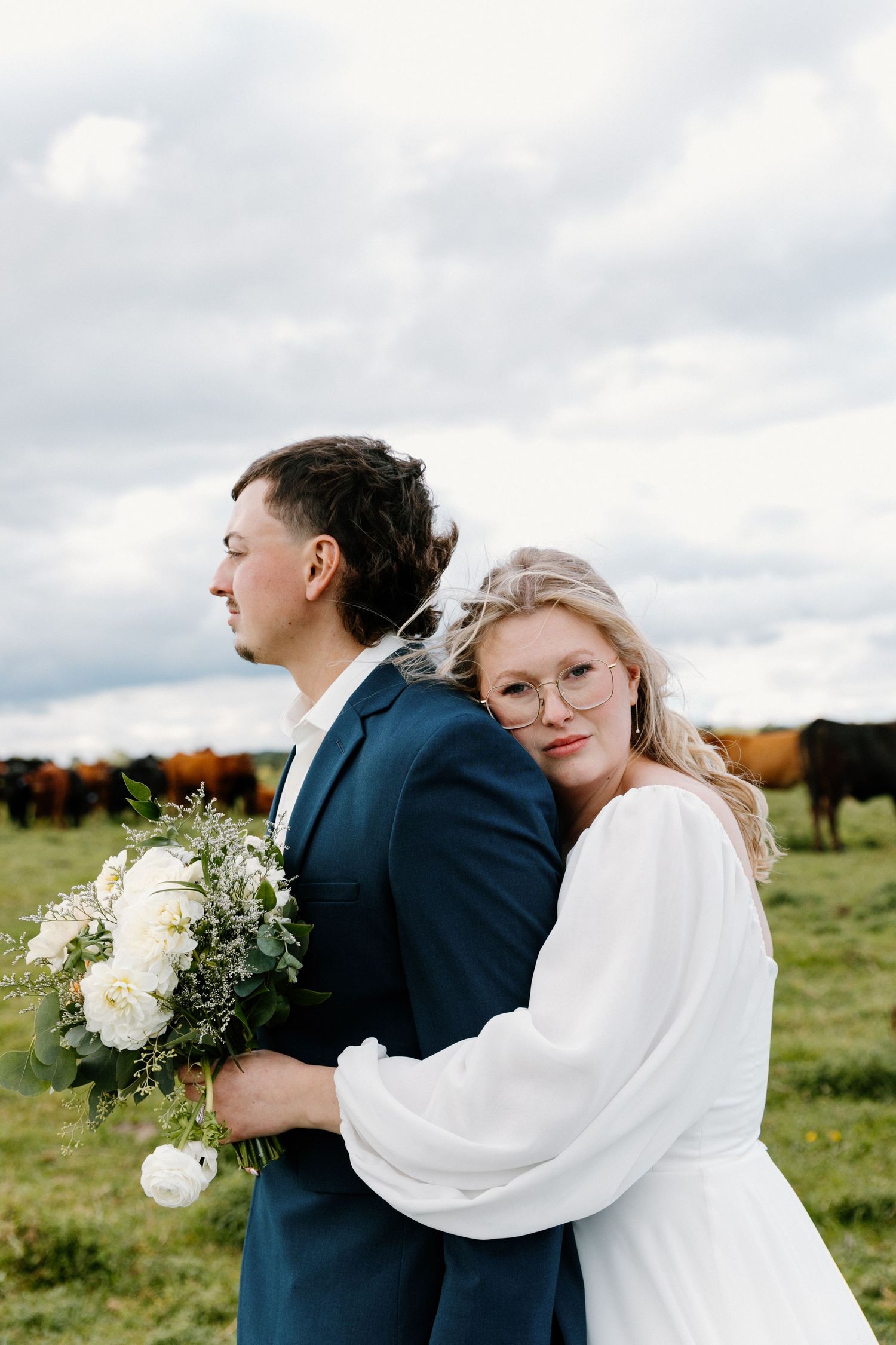 Bride in white dress with glasses embraces groom in navy suit, holding white floral bouquet in a green pasture.