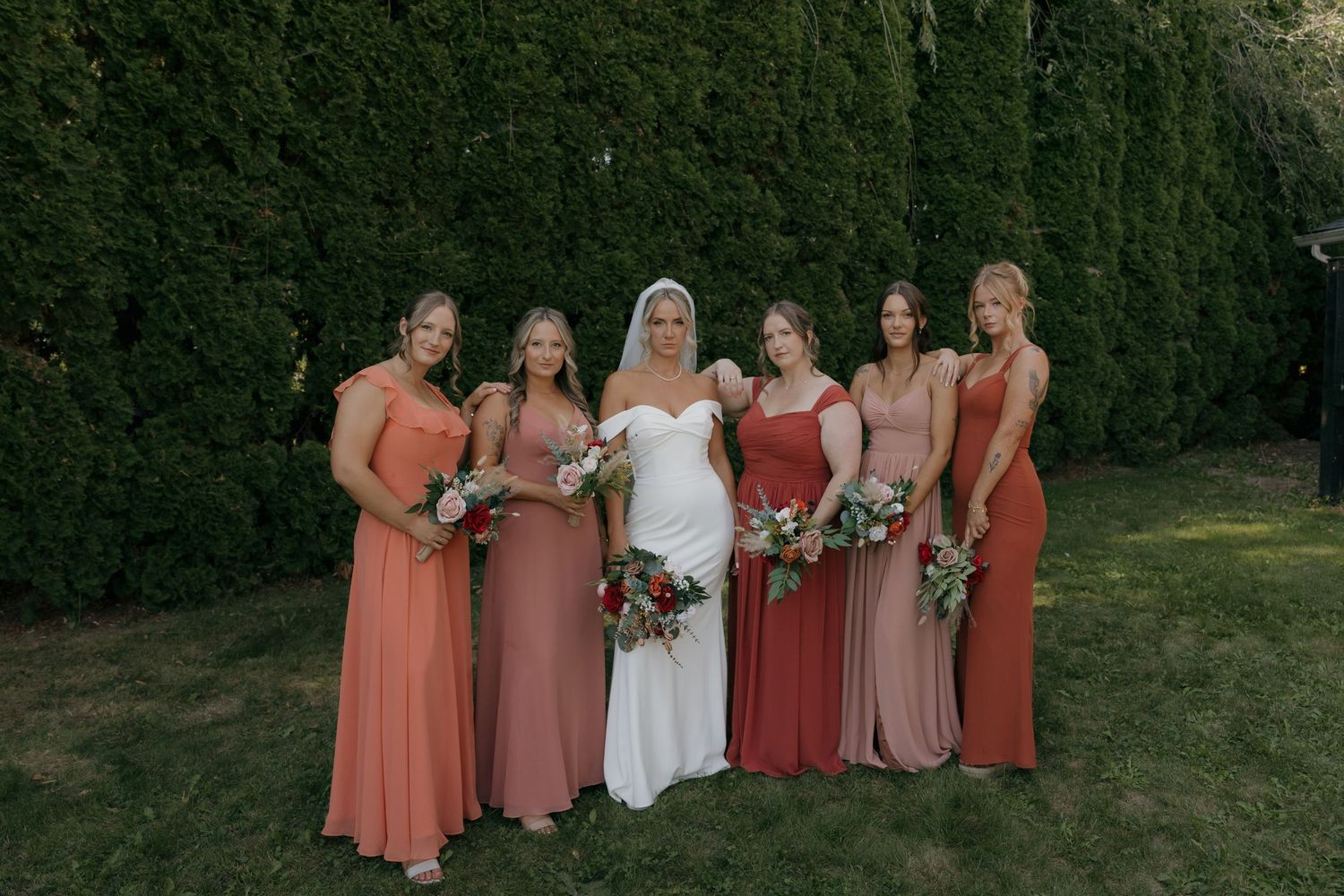 Bride with five bridesmaids in terracotta and mauve gowns holding bouquets before evergreen hedge.