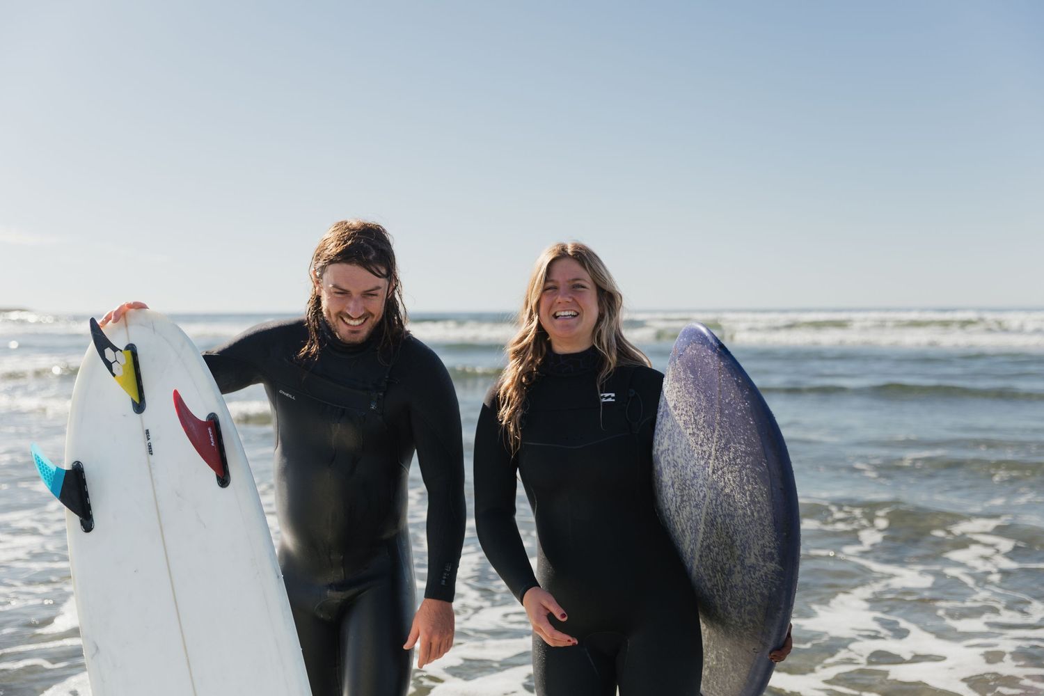 Two surfers in black wetsuits smiling and holding surfboards on a sunny beach.