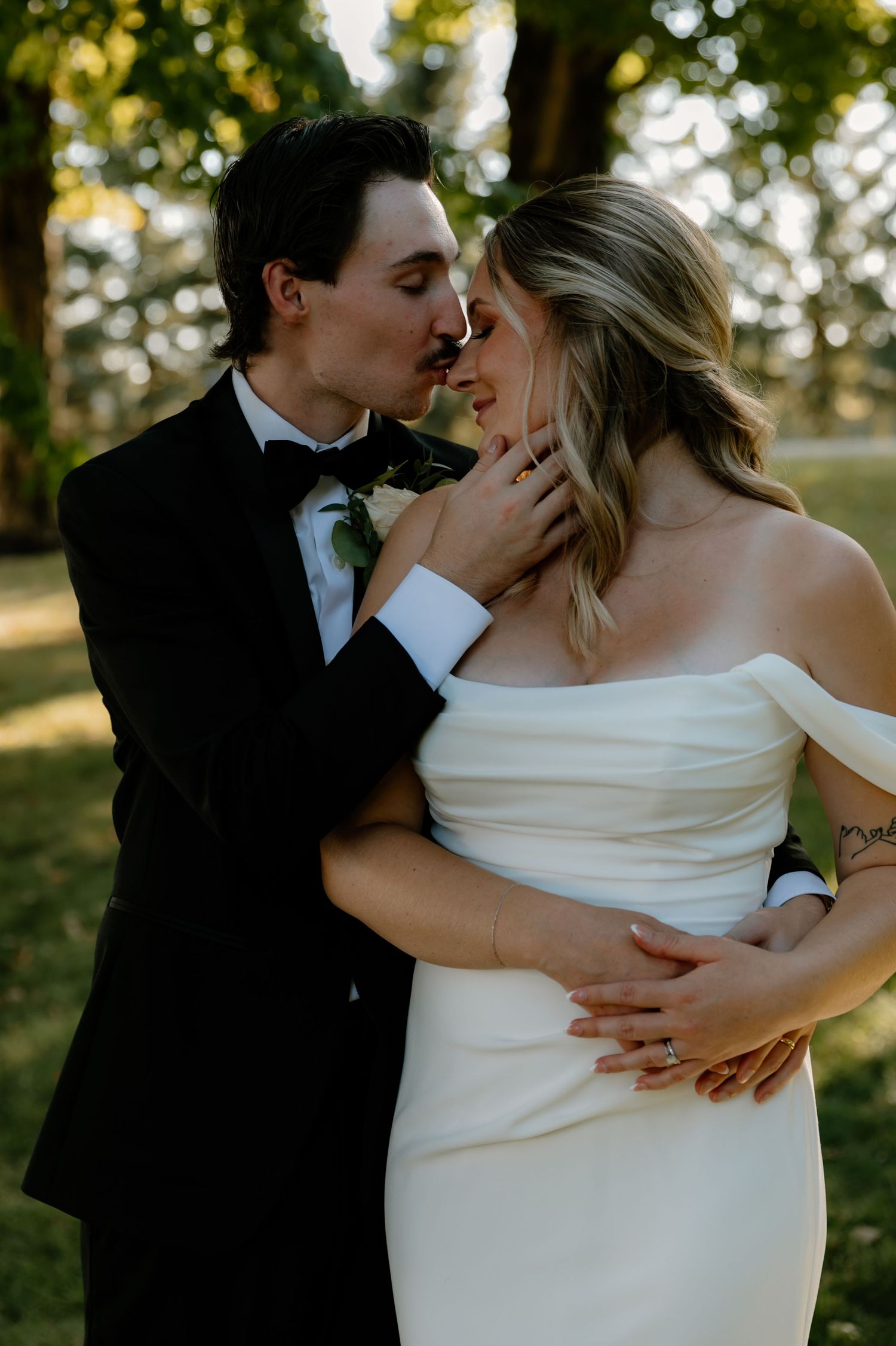 Bride and groom share an intimate moment, groom kissing bride's forehead in a sunlit outdoor wedding setting.