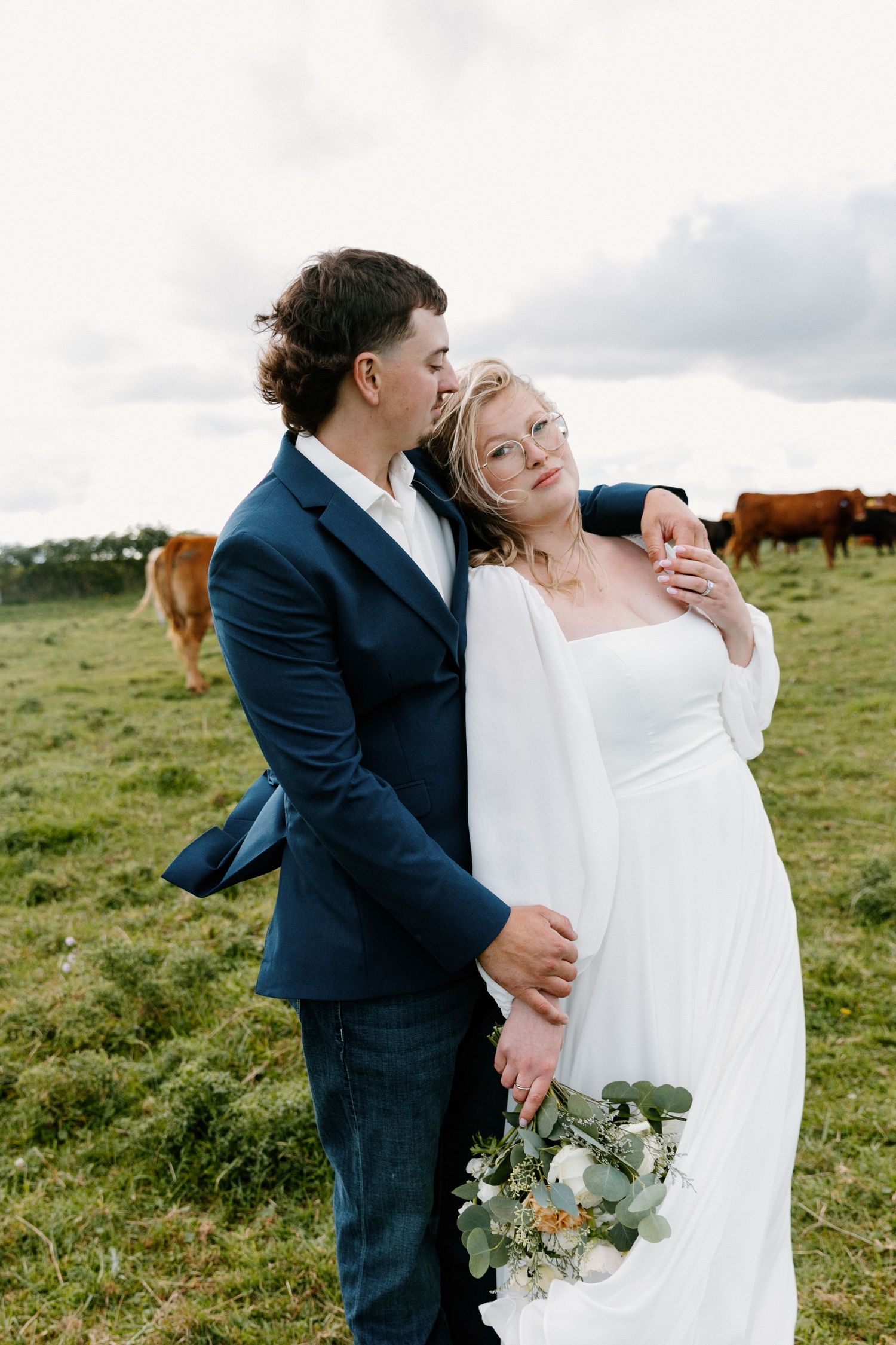 Bride and groom embrace in green pasture with cattle grazing in the background.