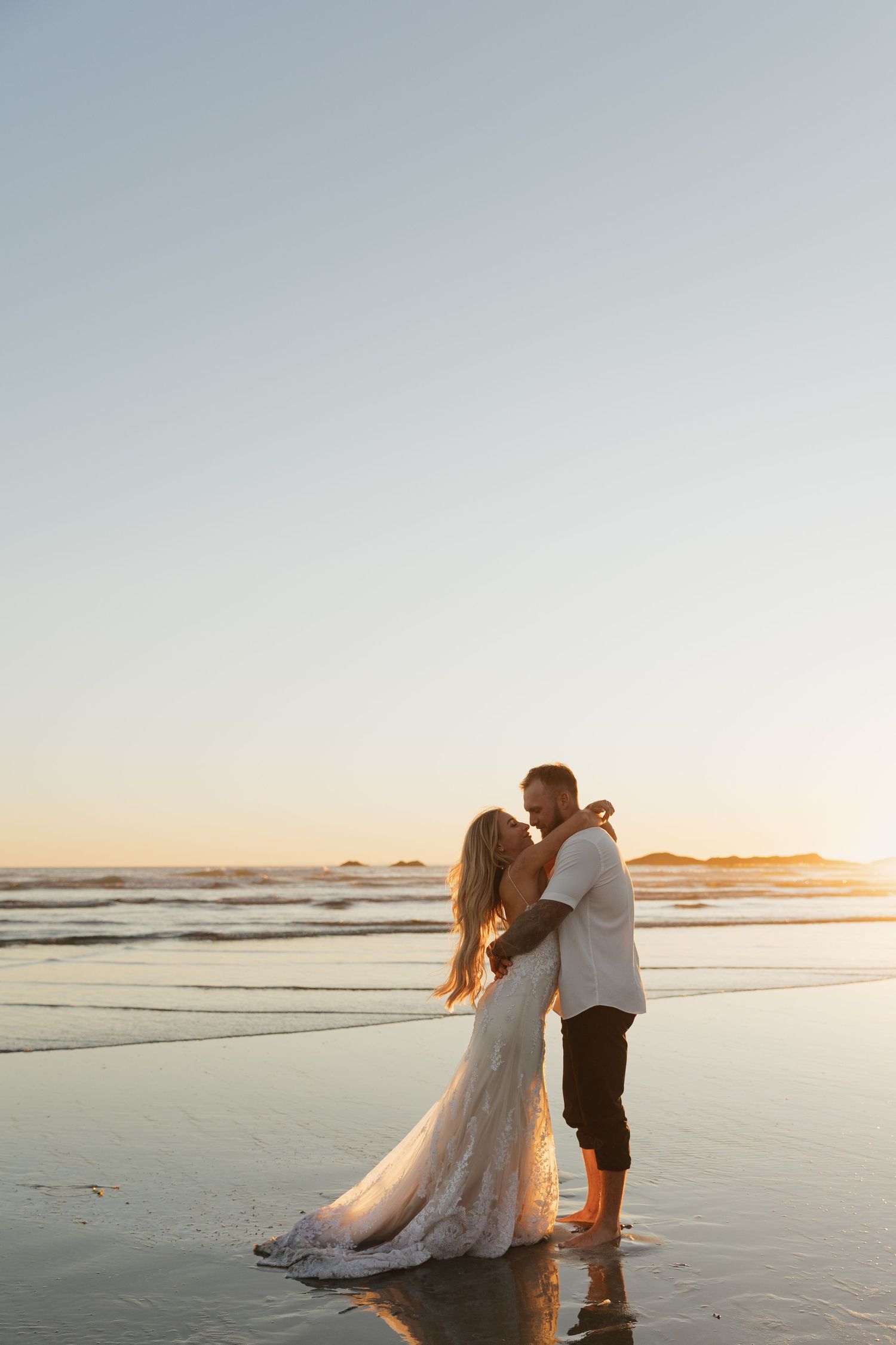 Newlyweds share a romantic kiss on a reflective beach at sunset, bride in flowing white lace gown.