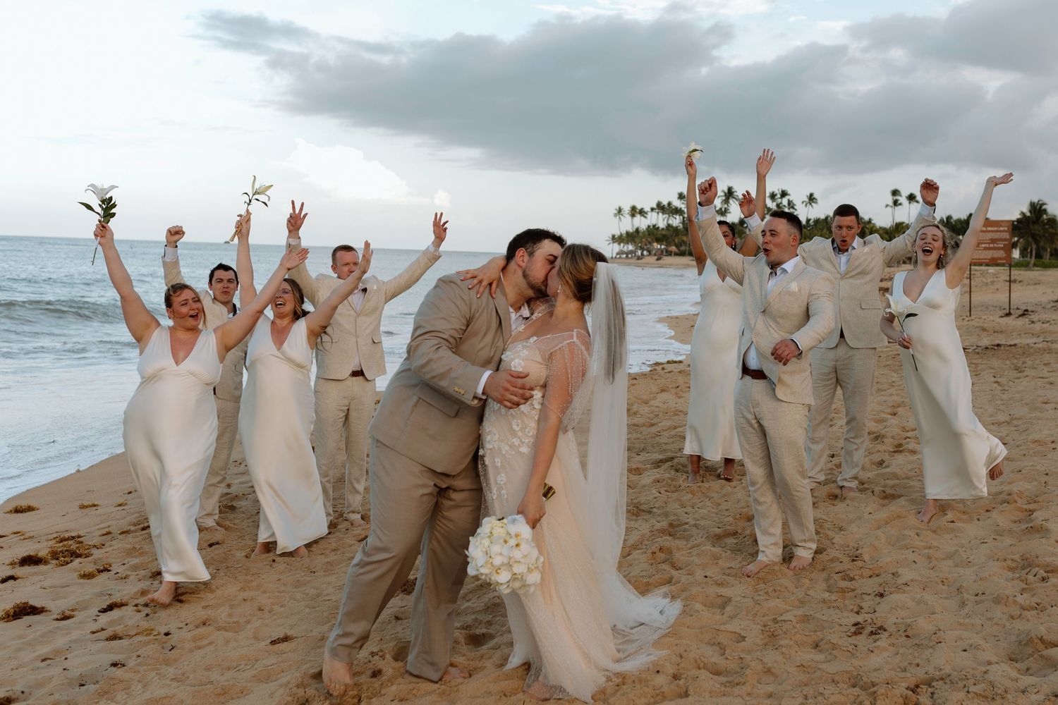 Bride and groom kiss surrounded by cheering wedding party on tropical beach.