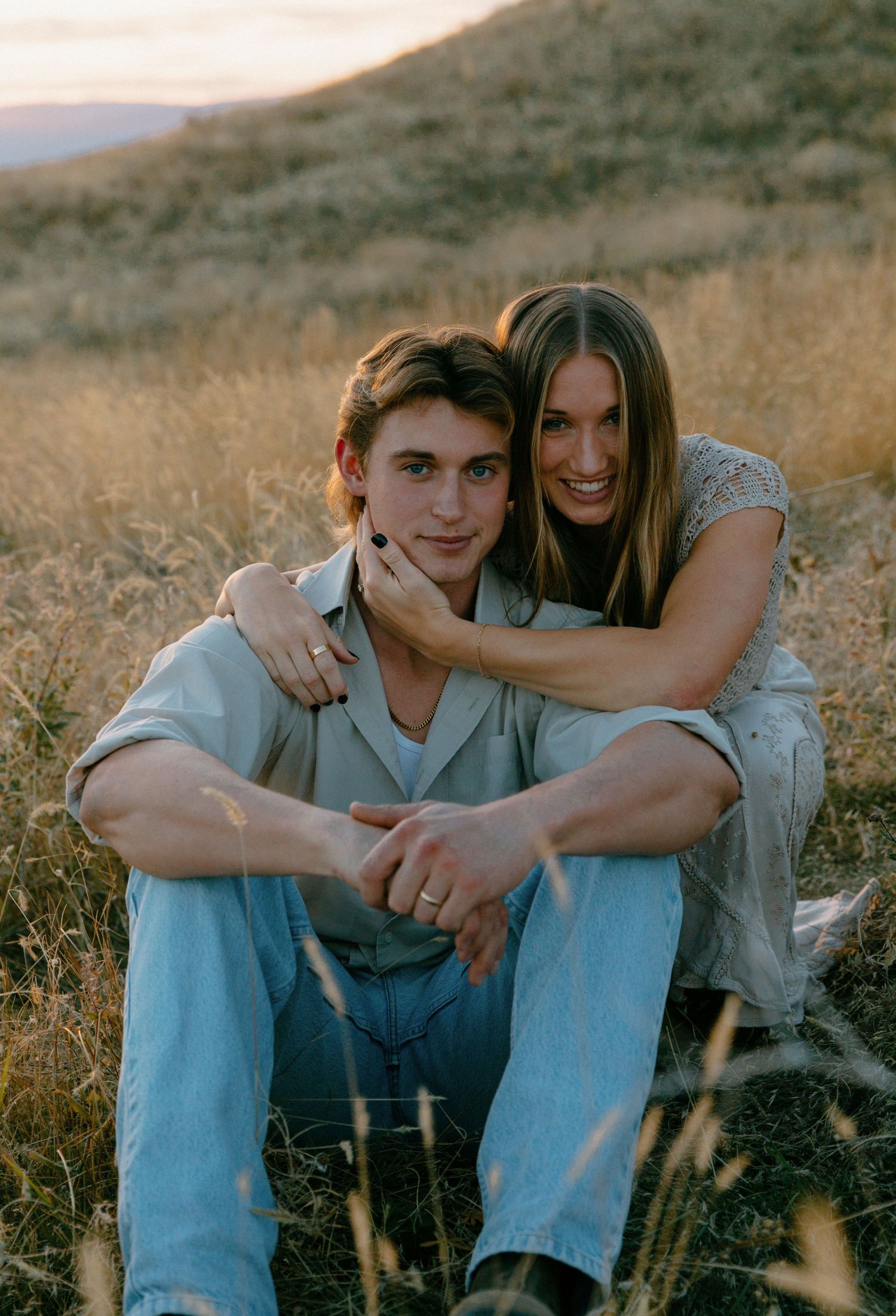Young couple sitting together in dry grass field during warm golden sunset.