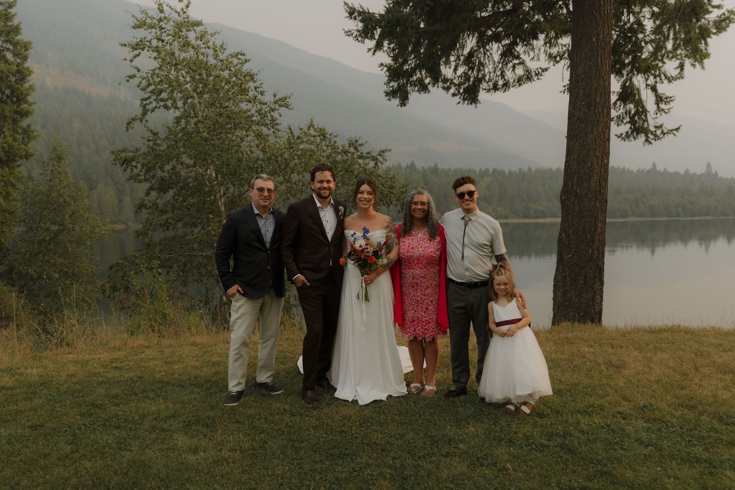 Wedding party family portrait by misty lake with bride, groom, and flower girl.