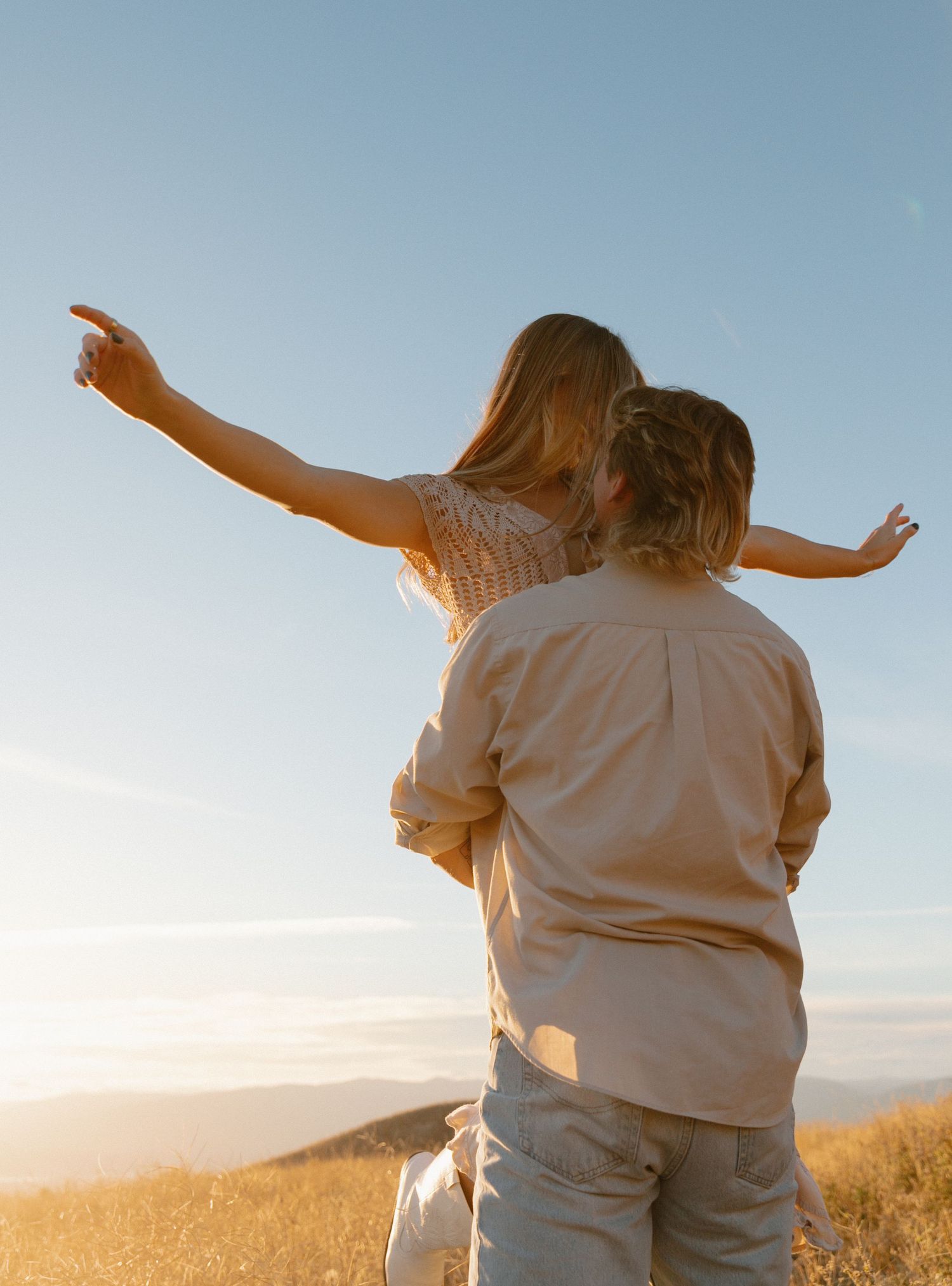 Couple standing on a hilltop at sunset, woman pointing toward the horizon with arms outstretched.