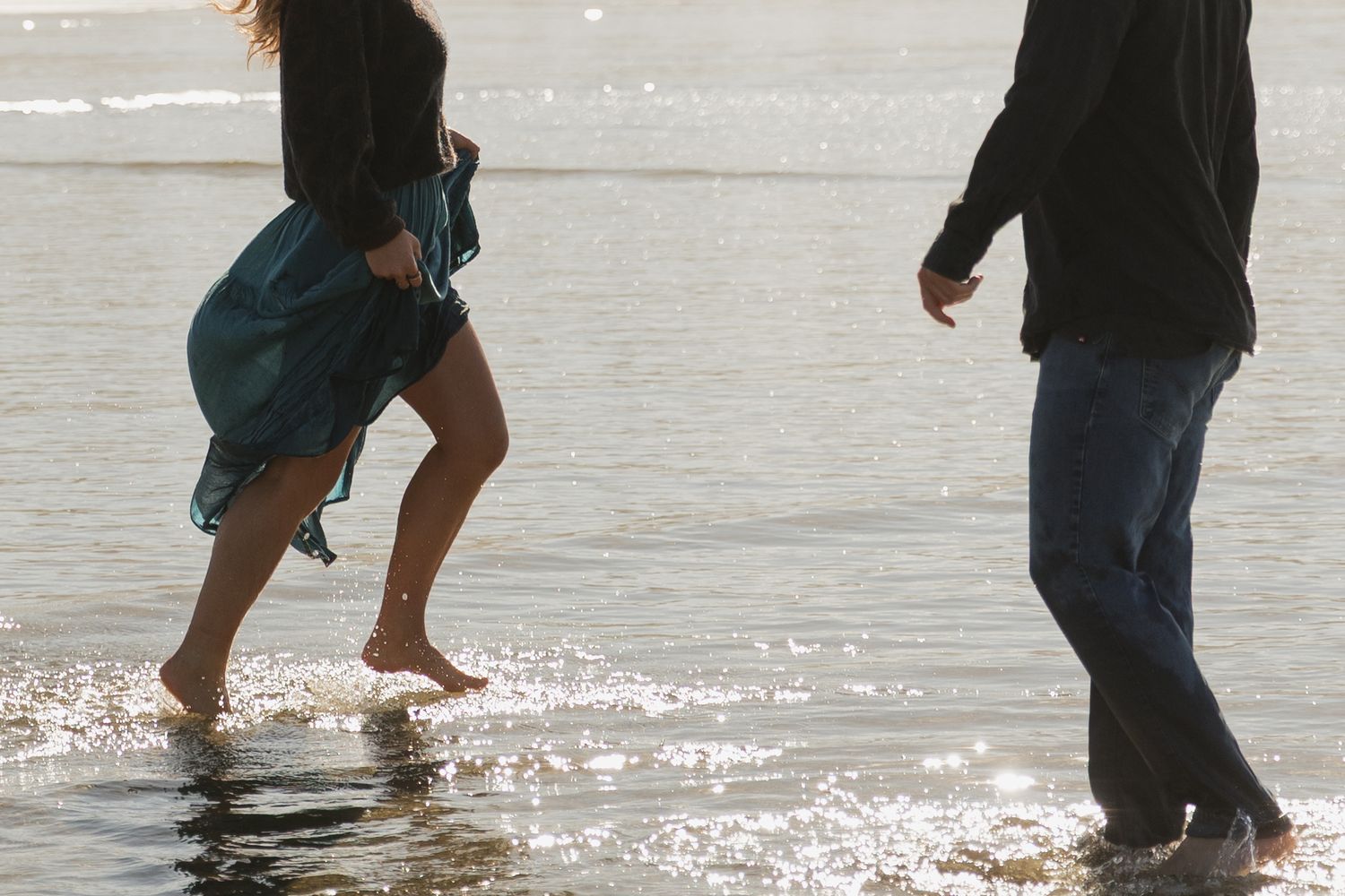 Couple walking barefoot through shallow ocean waves on a sunny beach, woman in teal skirt.