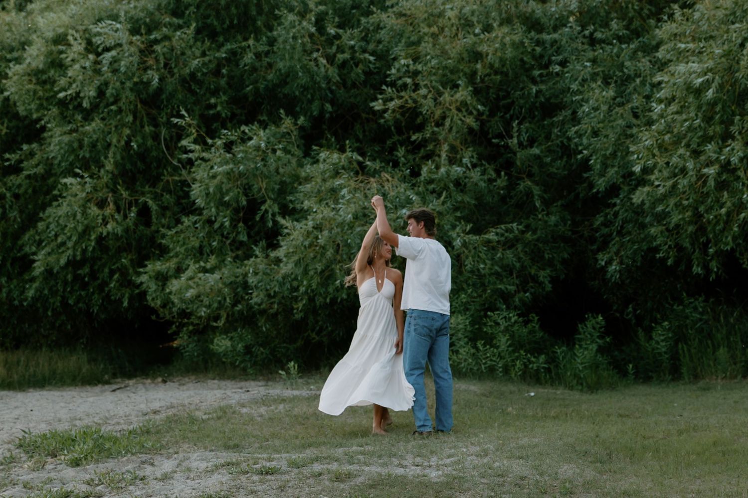 A couple dancing together in a grassy clearing surrounded by lush green trees in summer.