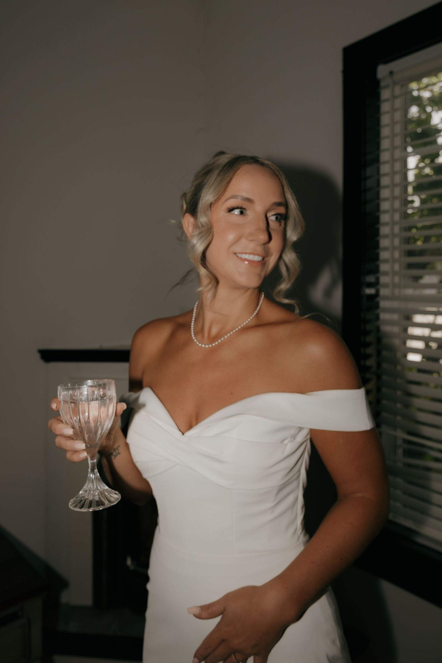 Smiling bride in off-shoulder white gown holding crystal champagne glass, wearing pearl necklace.