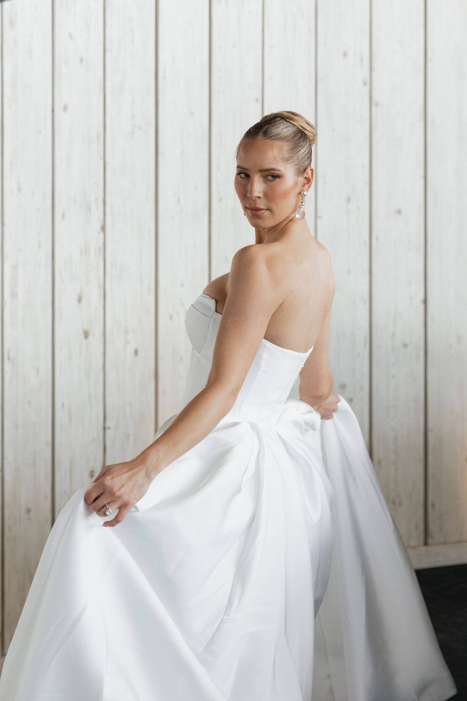 Elegant bride in strapless white ballgown with bow detail poses against light wood panel background.