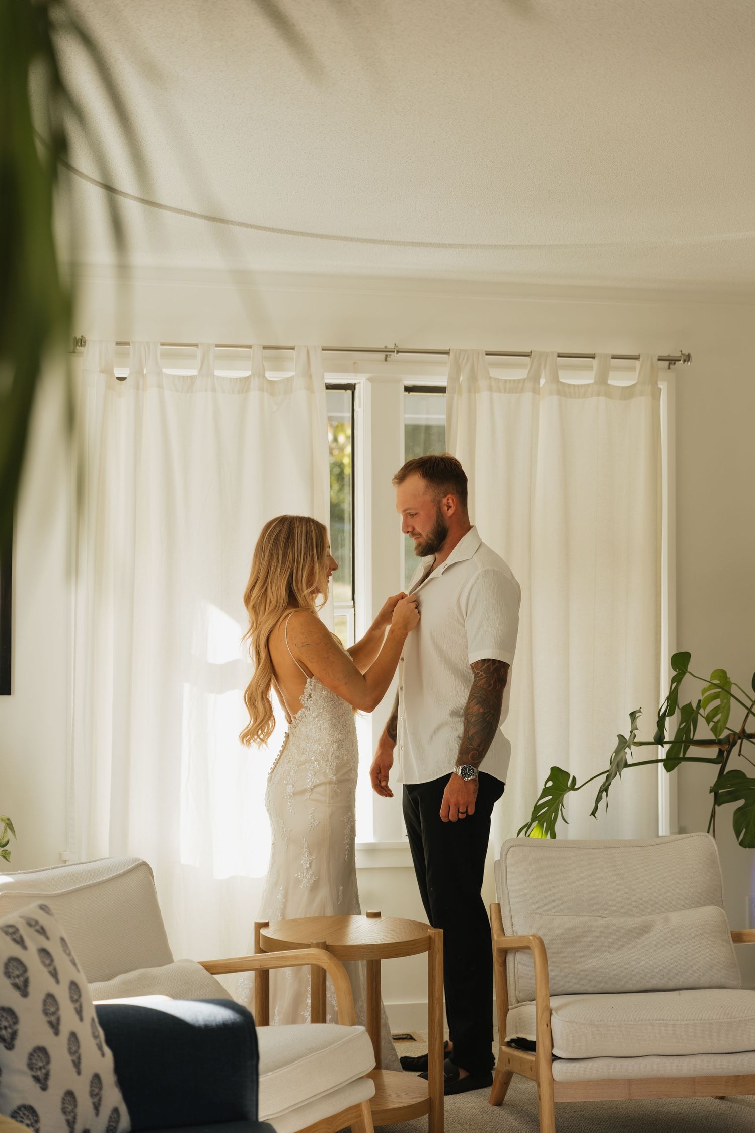 Bride in lace gown adjusts groom's collar in a bright, modern living room bathed in natural light.