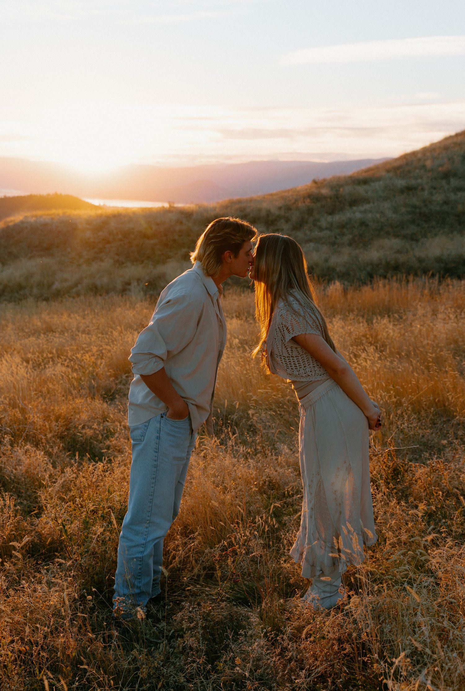 Couple sharing a kiss at golden sunset in a golden meadow with scenic mountain and lake views behind them.