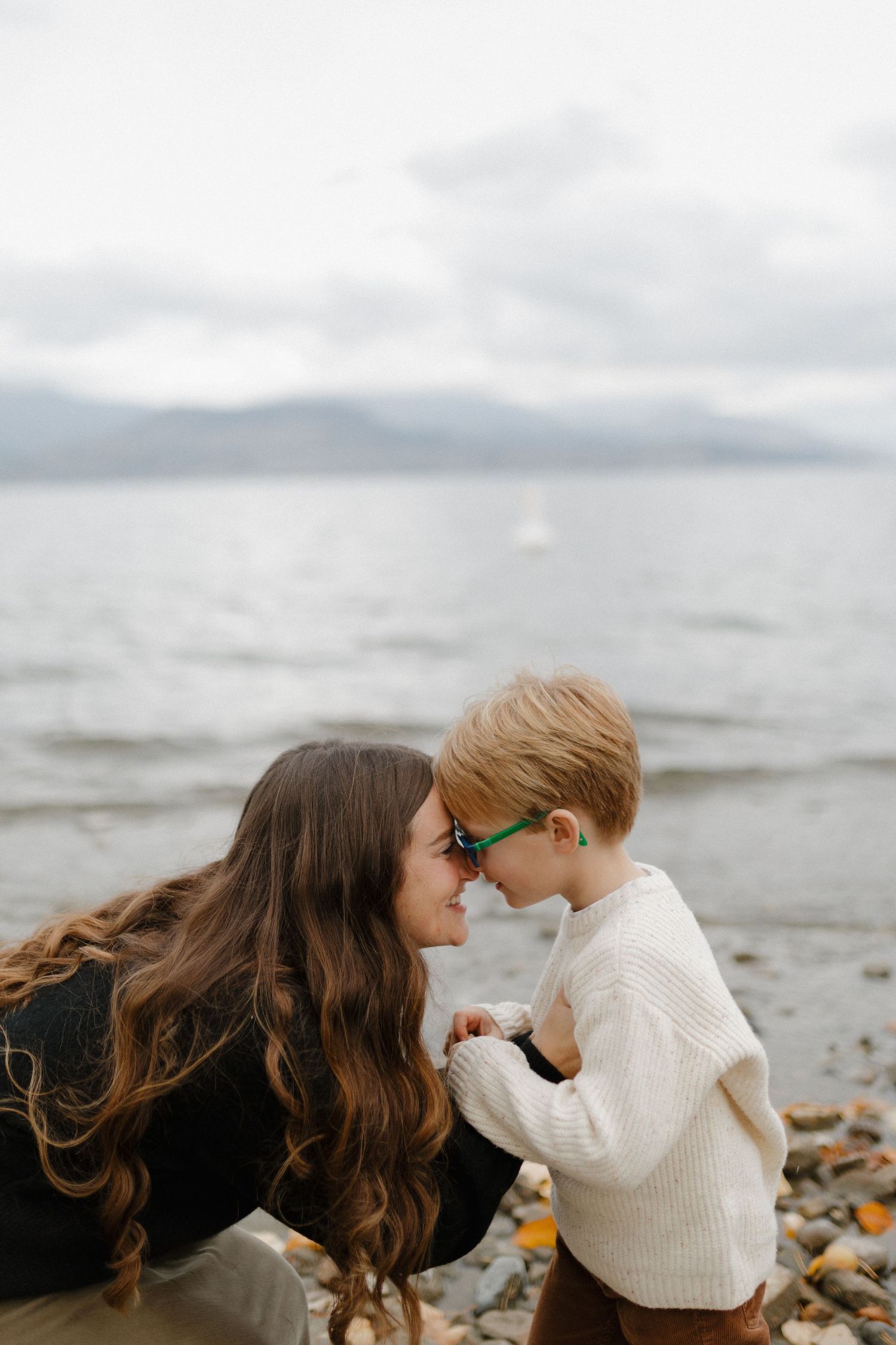 Mother and young boy touching foreheads affectionately on a rocky lakeside beach under cloudy skies.