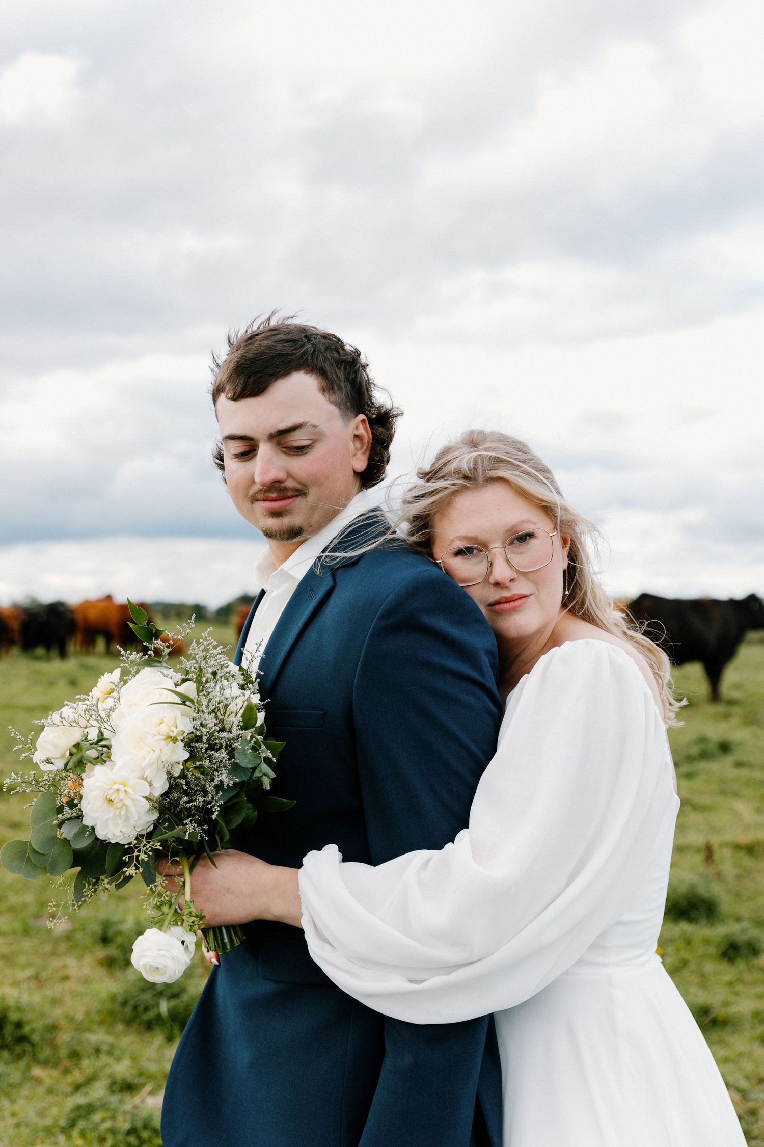 Bride hugs groom from behind holding white bouquet in a green pasture with cattle nearby.