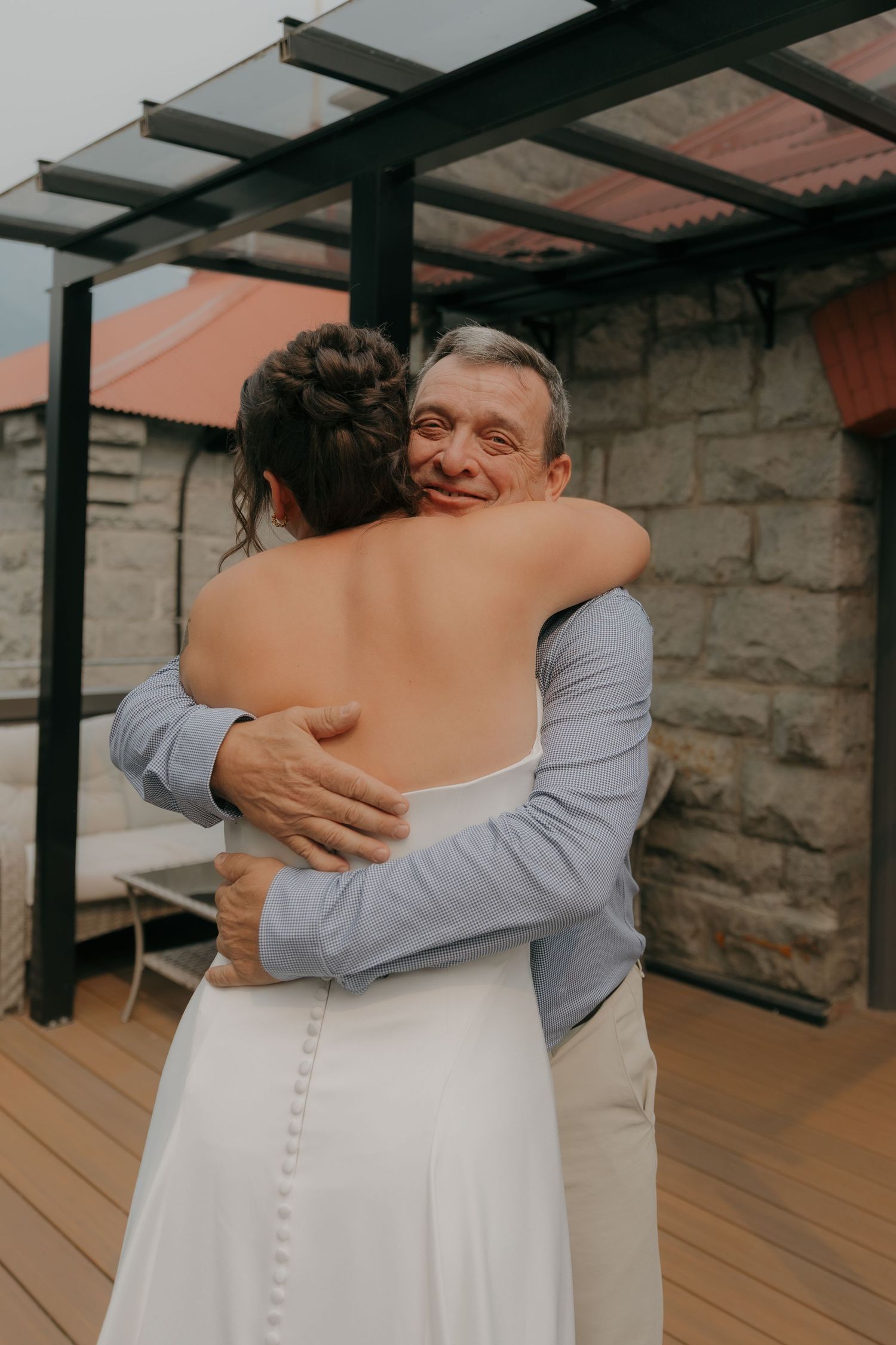 Emotional father hugs bride in backless white wedding dress on outdoor deck at reception.
