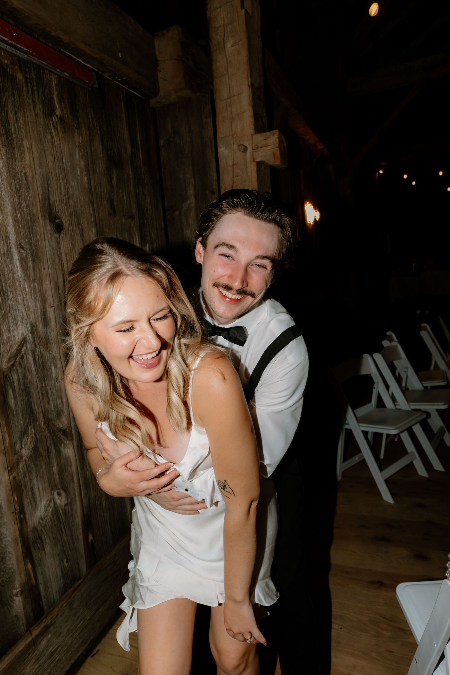 Joyful bride and groom laughing together in rustic barn wedding reception with string lights.