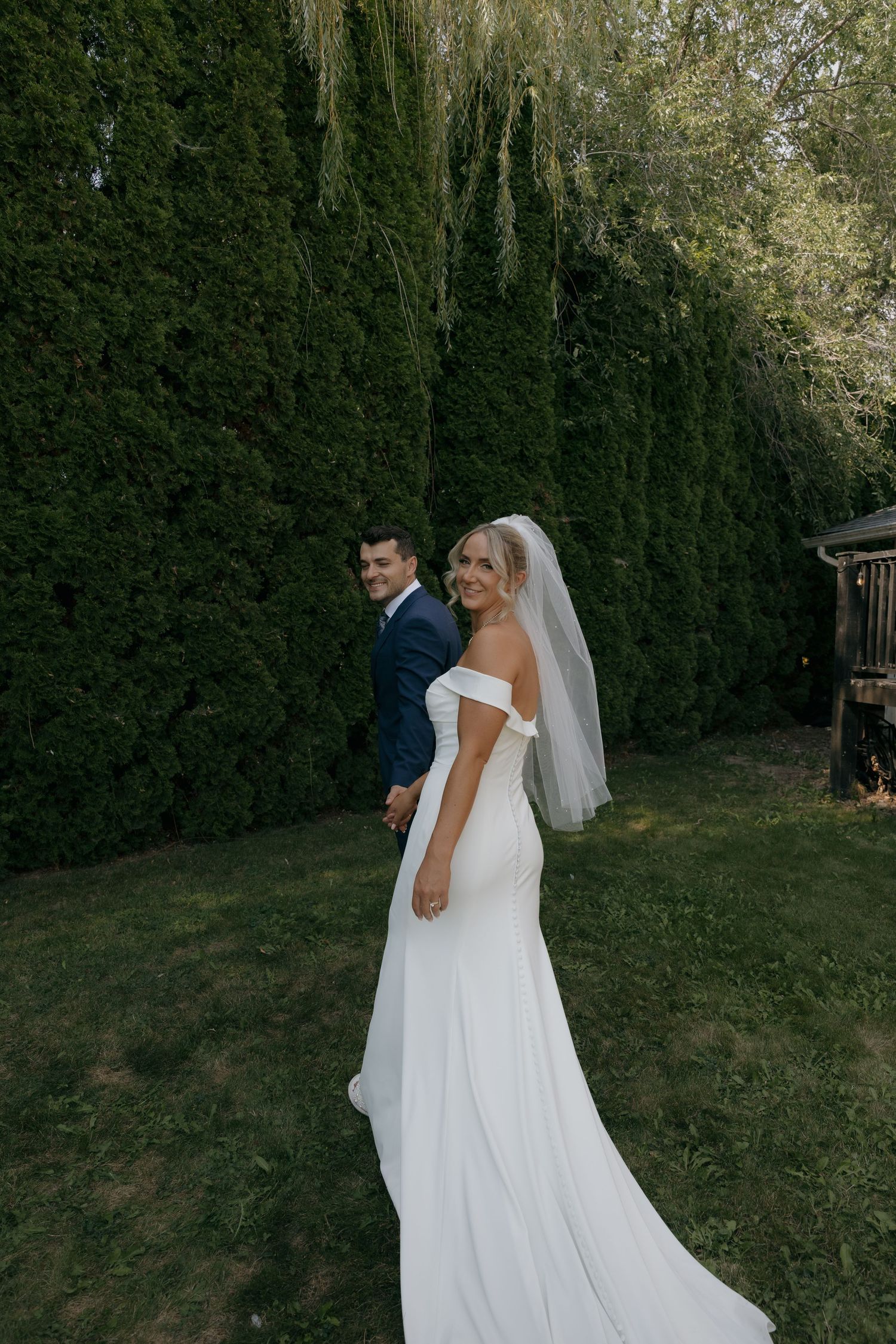 Bride in off-shoulder white gown with veil holds groom's hand outdoors near tall evergreen hedges.