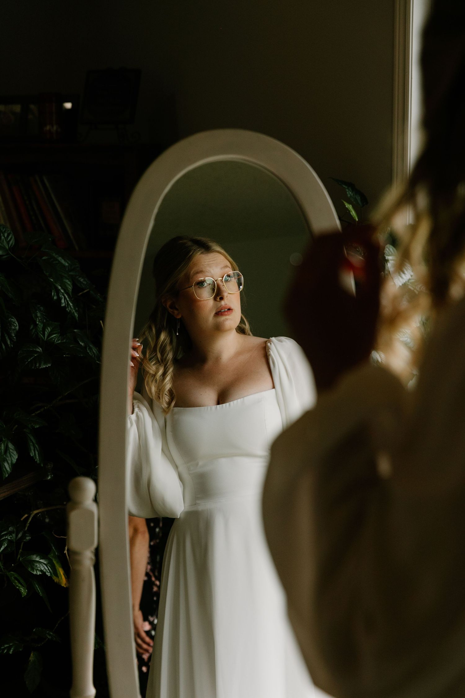 Bride in white square-neck gown with puff sleeves examines her reflection in an arched mirror.