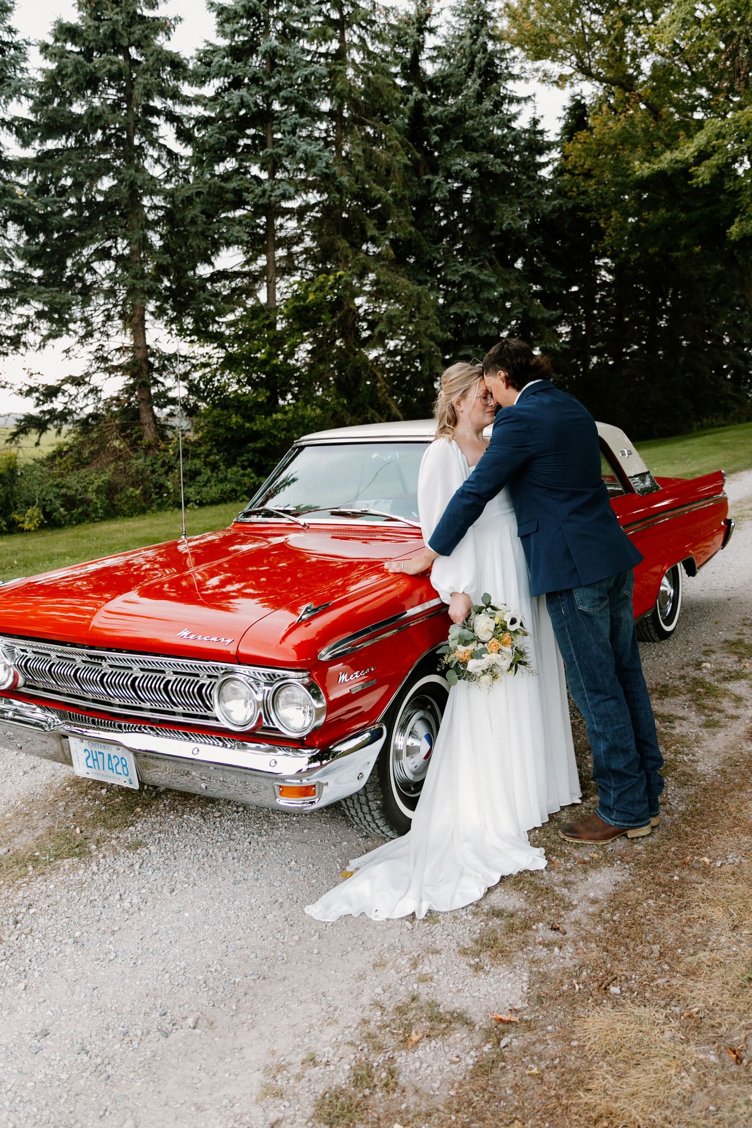 Wedding couple kissing beside a classic red vintage car surrounded by tall evergreen trees.